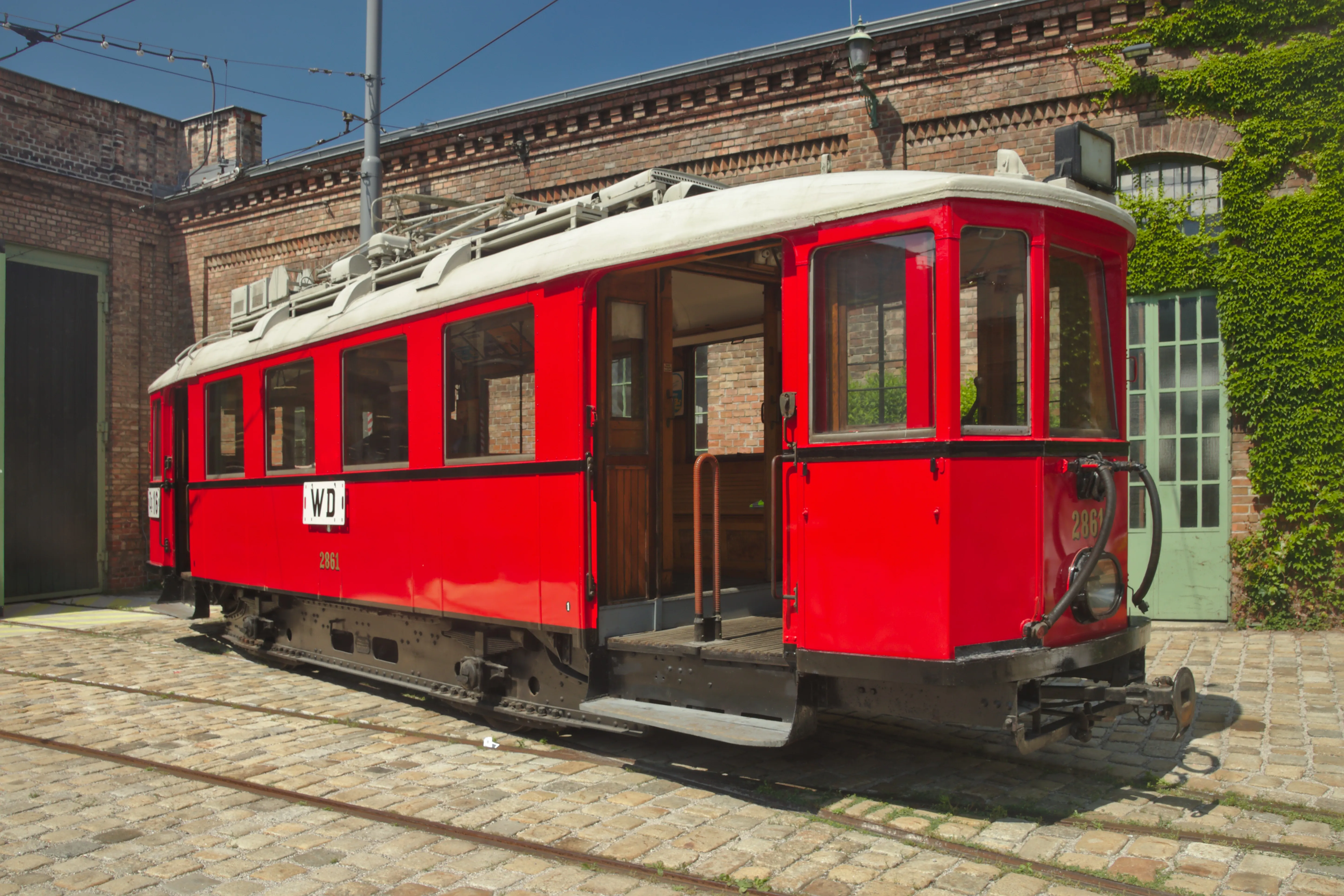 Czerwony zabytkowy tramwaj wystawiony przed ceglanym budynkiem z murami porośniętymi bluszczem