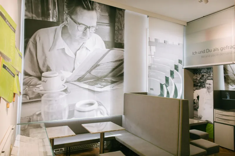 Interior of the Viktor Frankl Museum with a large mural of Viktor Frankl reading a newspaper and a seating area with tables and benches