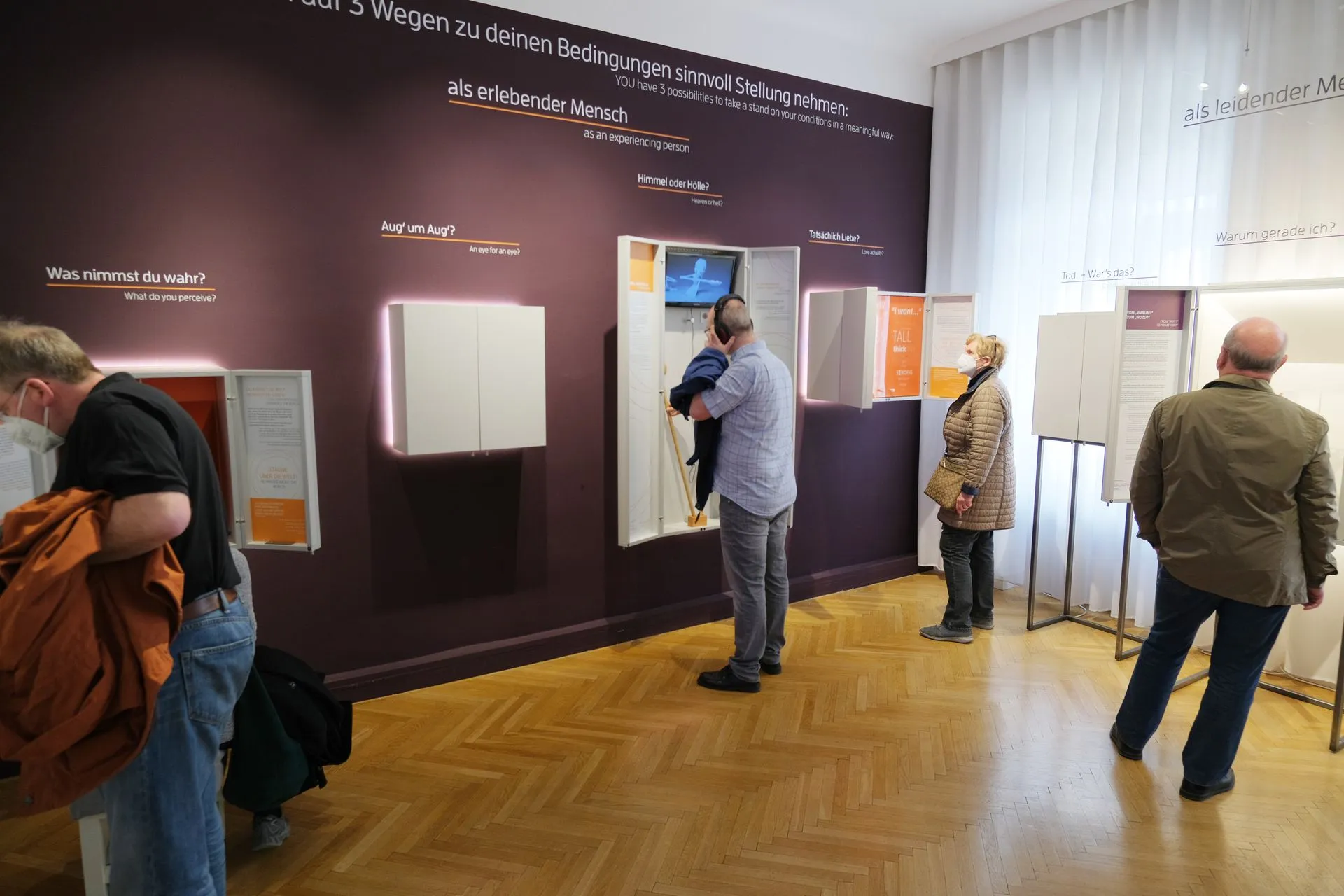 Visitantes viendo paneles de exhibición y pantallas interactivas en una galería de museo con suelo de madera y paredes moradas