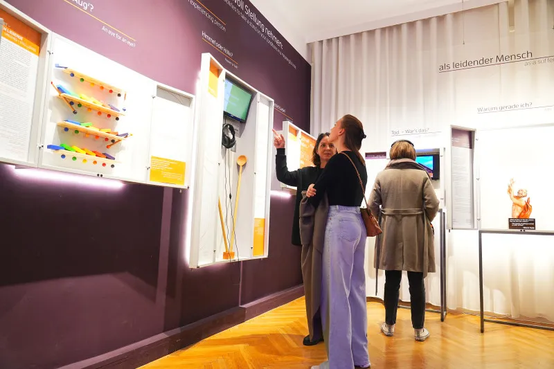 Dos mujeres interactuando con una exposición en una galería de museo con paredes moradas y vitrinas