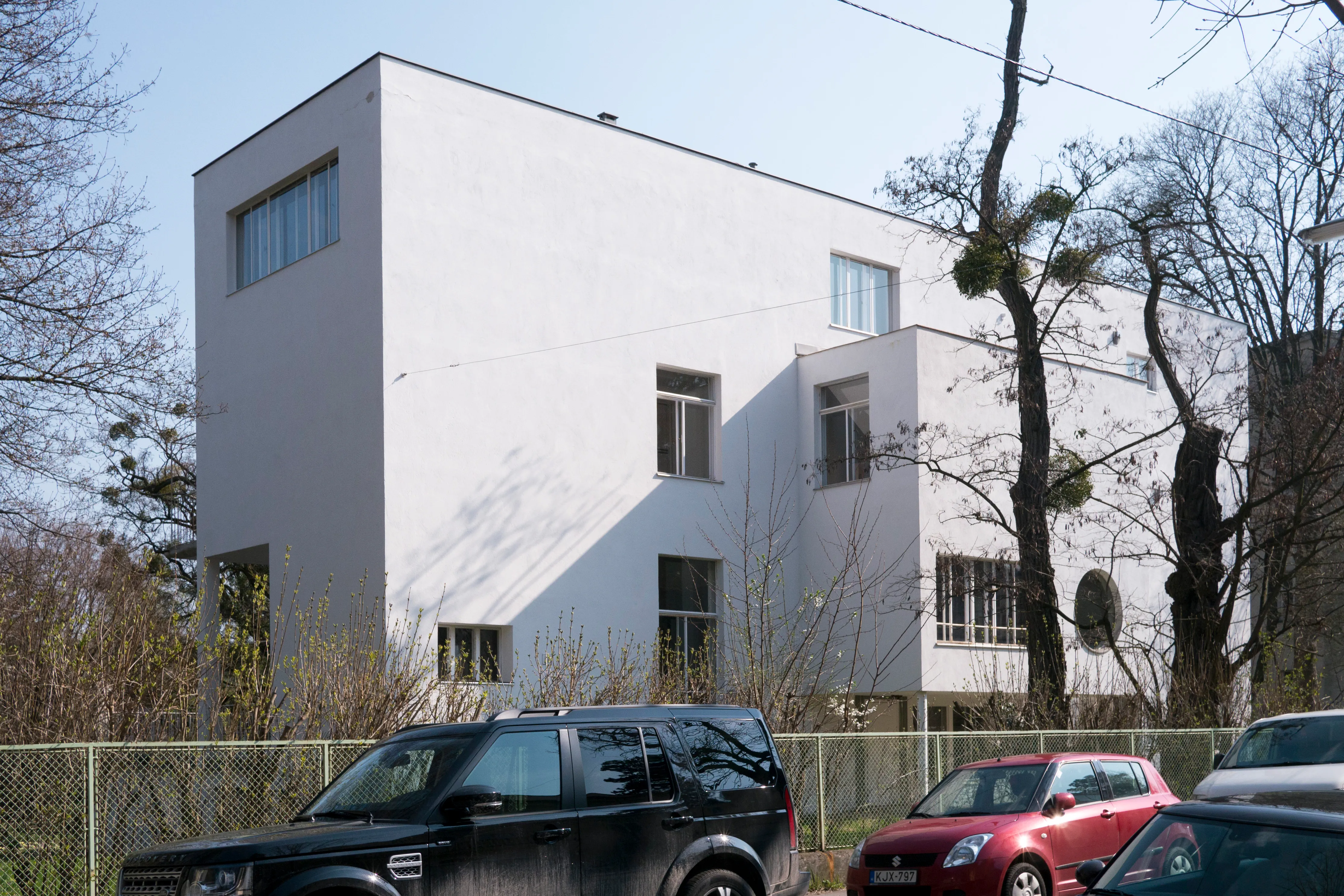 Un edificio modernista blanco con ventanas rectangulares y techo plano, visto desde el frente con árboles y coches en primer plano.