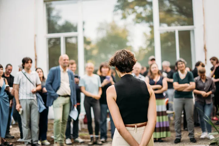 Una persona con pelo corto y top negro está de espaldas a la cámara, dirigiéndose a un grupo diverso de personas frente a un edificio moderno con grandes ventanales.