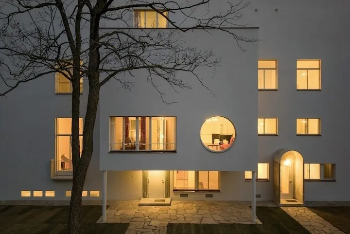 Edificio modernista blanco con ventanas rectangulares y circulares iluminadas por la noche, un árbol desnudo en primer plano