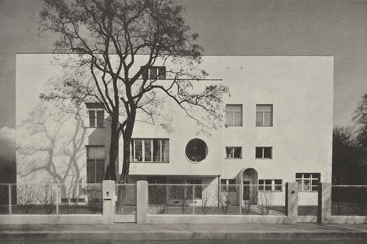 Un edificio modernista blanco con una ventana circular, ventanas rectangulares y un árbol delante.