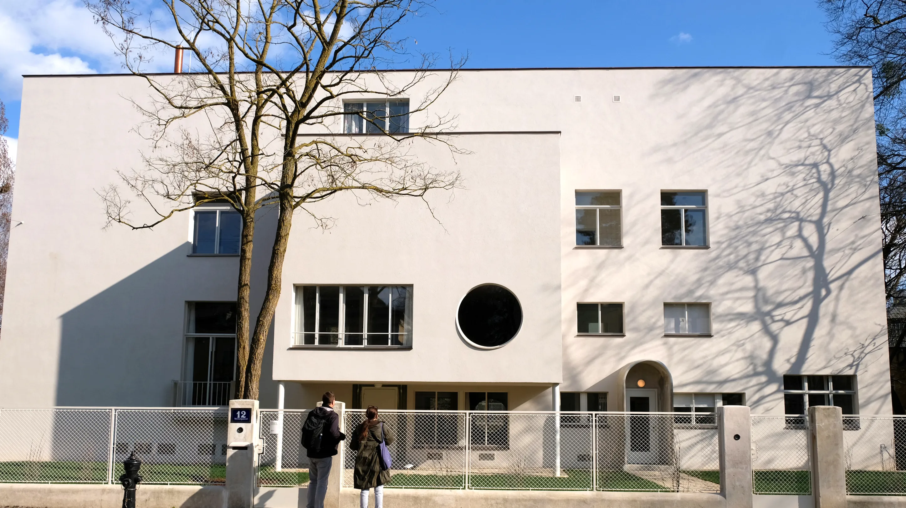 Un edificio modernista blanco con una ventana circular y ventanas rectangulares, un árbol con ramas desnudas que proyecta sombras y dos personas cerca de la entrada.