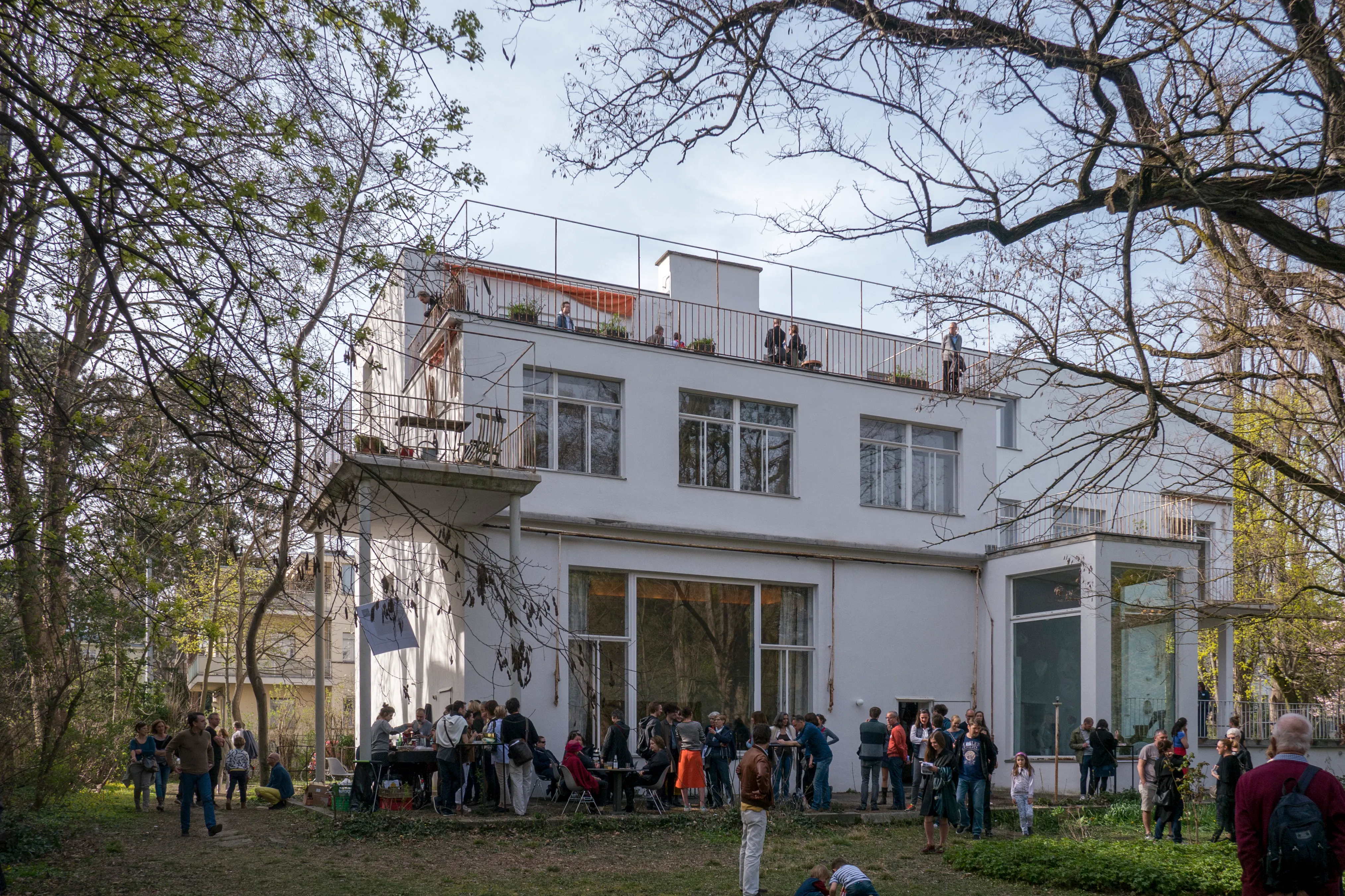 Vista exterior de la Villa Beer, de estilo modernista y color blanco, con personas reunidas en el jardín y en la terraza de la azotea.