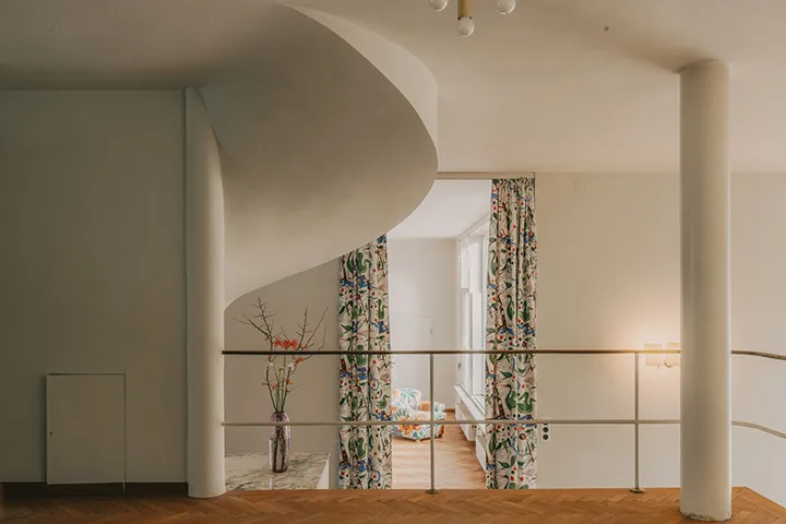 vista interior de Villa Beer con una escalera blanca curva, cortinas florales y suelos de madera