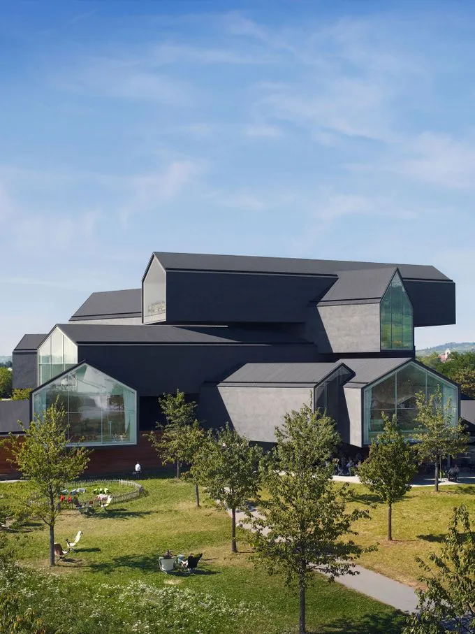 Ciemne, kanciaste i spiętrzone bryły architektoniczne ze szklanymi szczytami, wznoszące się nad zielonym trawnikiem z młodymi drzewami pod błękitnym niebem