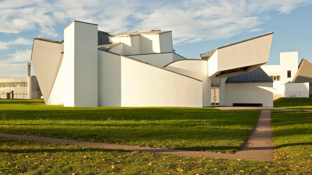 Wit hoekig deconstructivistisch museumgebouw met scherpe geometrische vormen, een groen gazon en een betonnen looppad onder een blauwe lucht met verspreide wolken