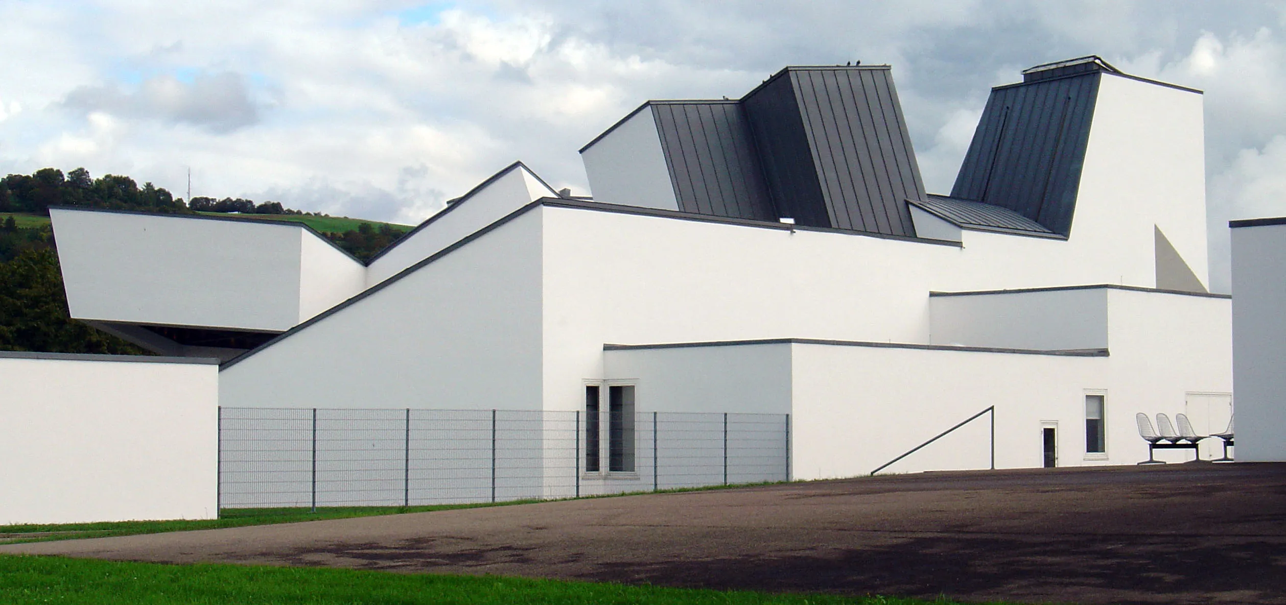 Wit hoekig deconstructivistisch gebouw met donkergrijze schuine daksneden, metalen hek, verharde binnenplaats en bewolkte lucht — het Vitra Design Museum