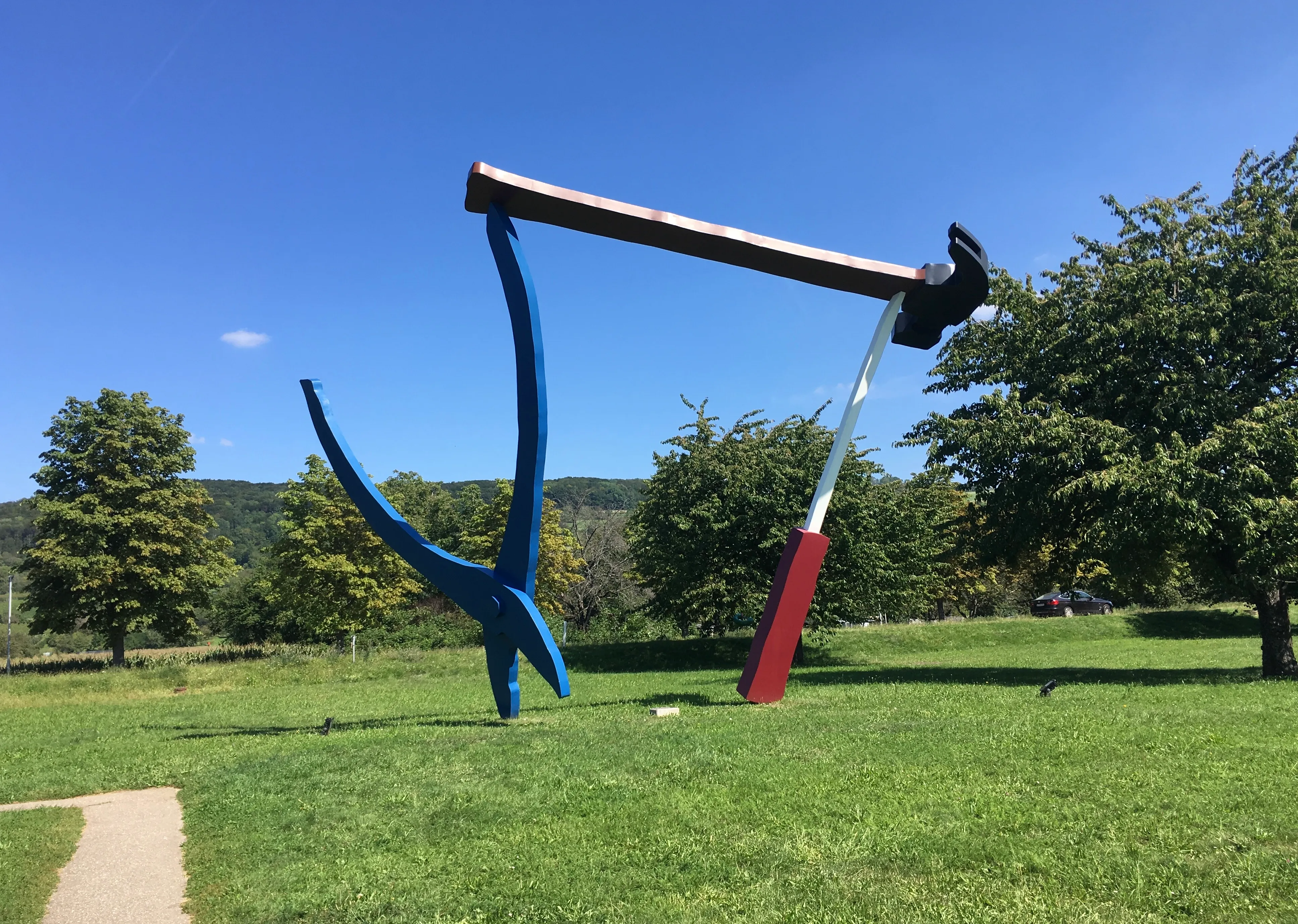 Groot buitenbeeld van overmaatse balancerende gereedschappen, waaronder een blauwe koevoet, een hamer met een rode steel en een houten balk op een grasveld onder een blauwe lucht