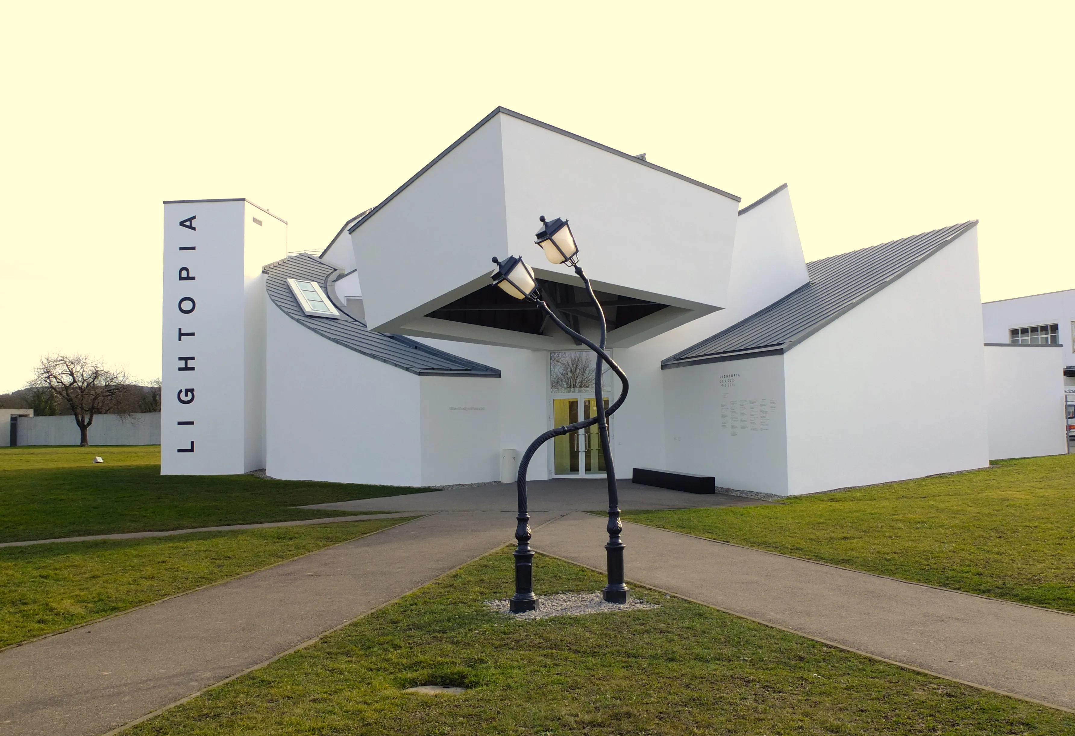 Buitenaanzicht van het witte, hoekige Vitra Design Museum-gebouw met een verticaal 'LIGHTOPIA'-bord en een gebogen zwarte lantaarnpaal op de voorgrond op een grasveld.