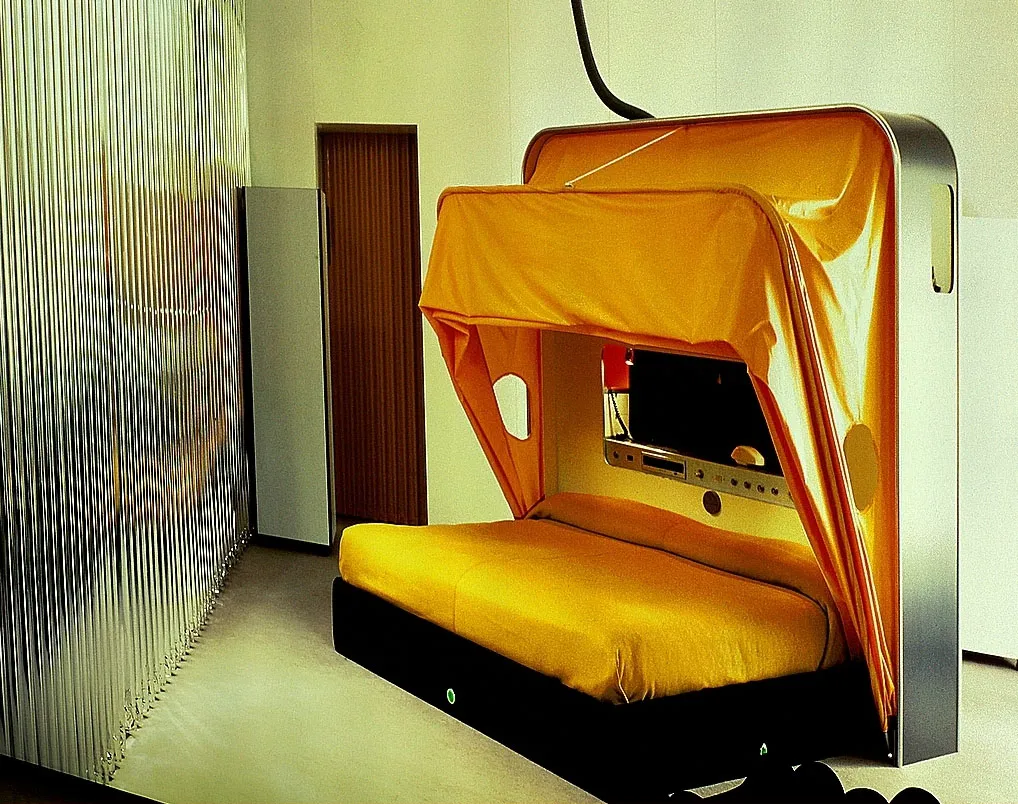Een retro-futuristisch cabrioletbed met een oranje stoffen kap en geel matras opgesteld in de tentoonstelling, voorzien van een zilveren metalen frame met een ingebouwd bedieningspaneel, geplaatst tegen een muur van golfplaat.