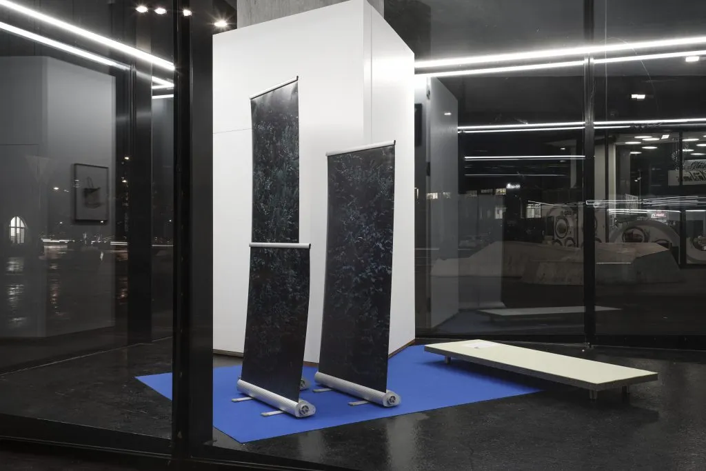 Ciemne pionowe panele na metalowych stojakach ustawione na niebieskiej platformie wewnątrz galerii ze szklanymi ścianami, obok której znajduje się biała ławka