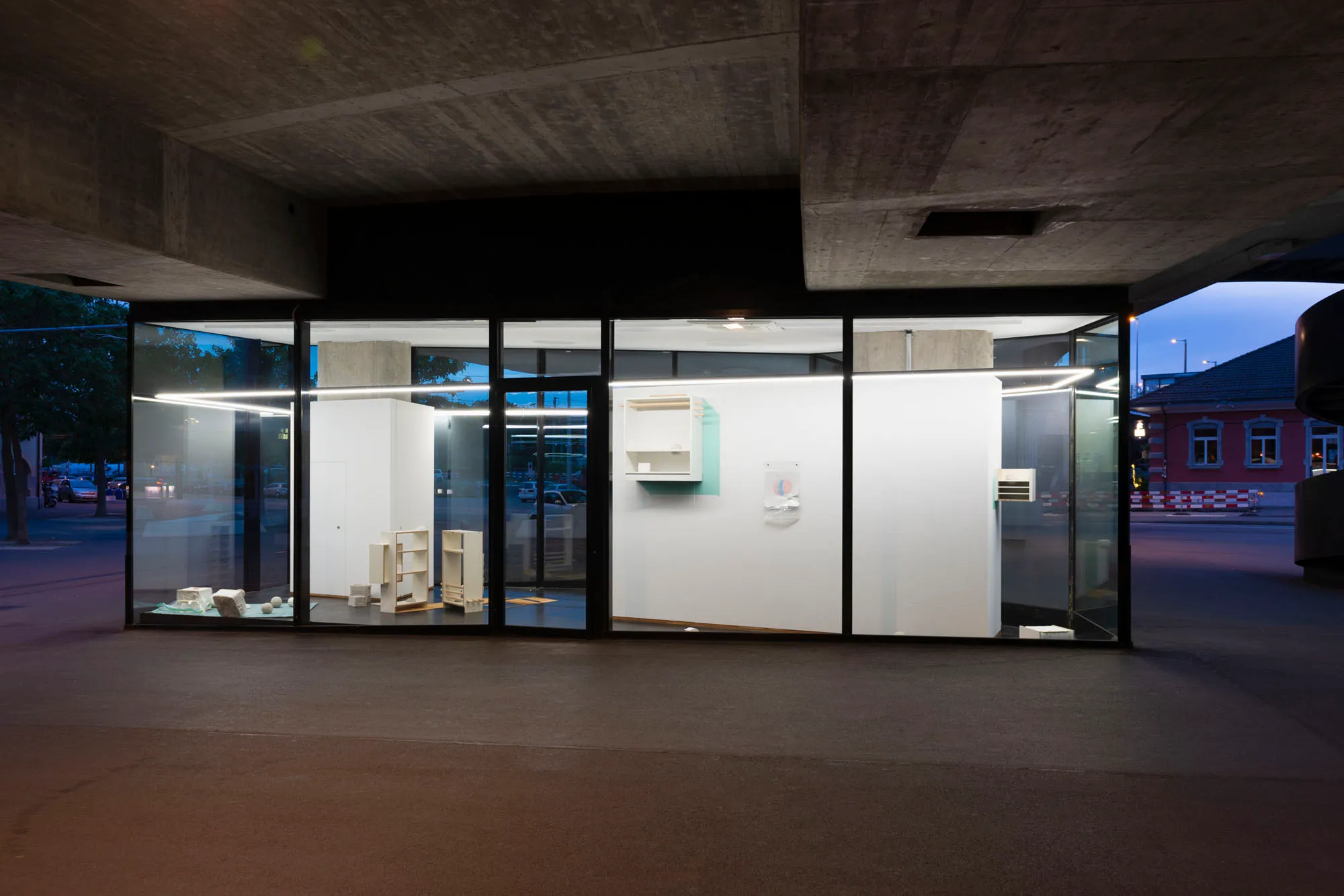 Przeszklona przestrzeń galerii, oświetlona nocą pod betonowym wiaduktem, z widocznymi przez przezroczystą fasadę białymi ścianami i dziełami sztuki.