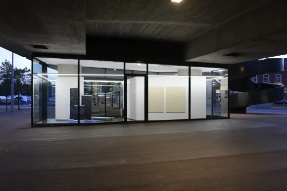 Przeszklona galeria oświetlona o zmierzchu pod betonowym wiaduktem, z dziełami sztuki widocznymi wewnątrz przezroczystej sali wystawowej
