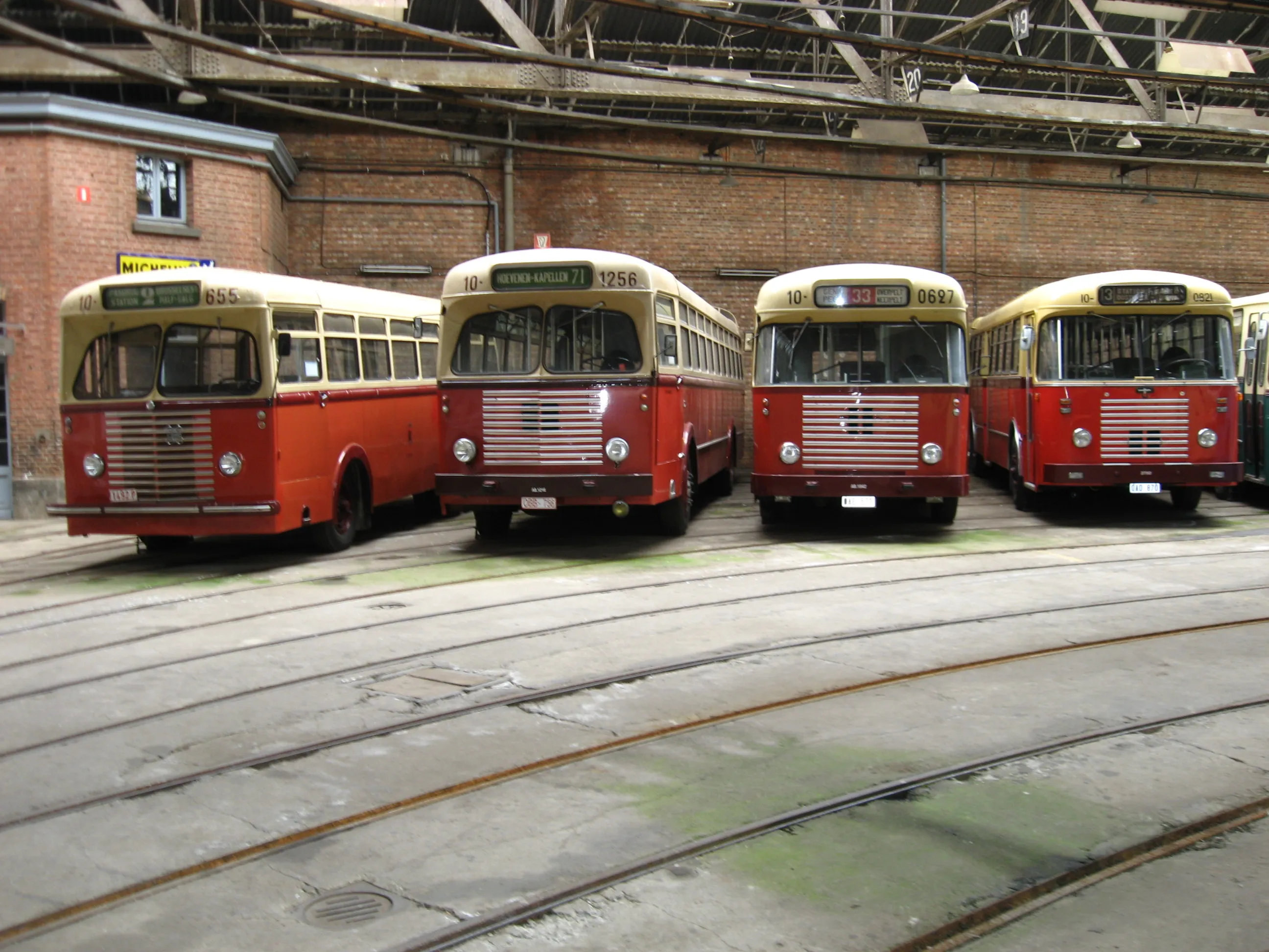 Cuatro autobuses antiguos de color rojo y crema aparcados uno al lado del otro dentro de un depósito de ladrillo con vías de tranvía que atraviesan el suelo