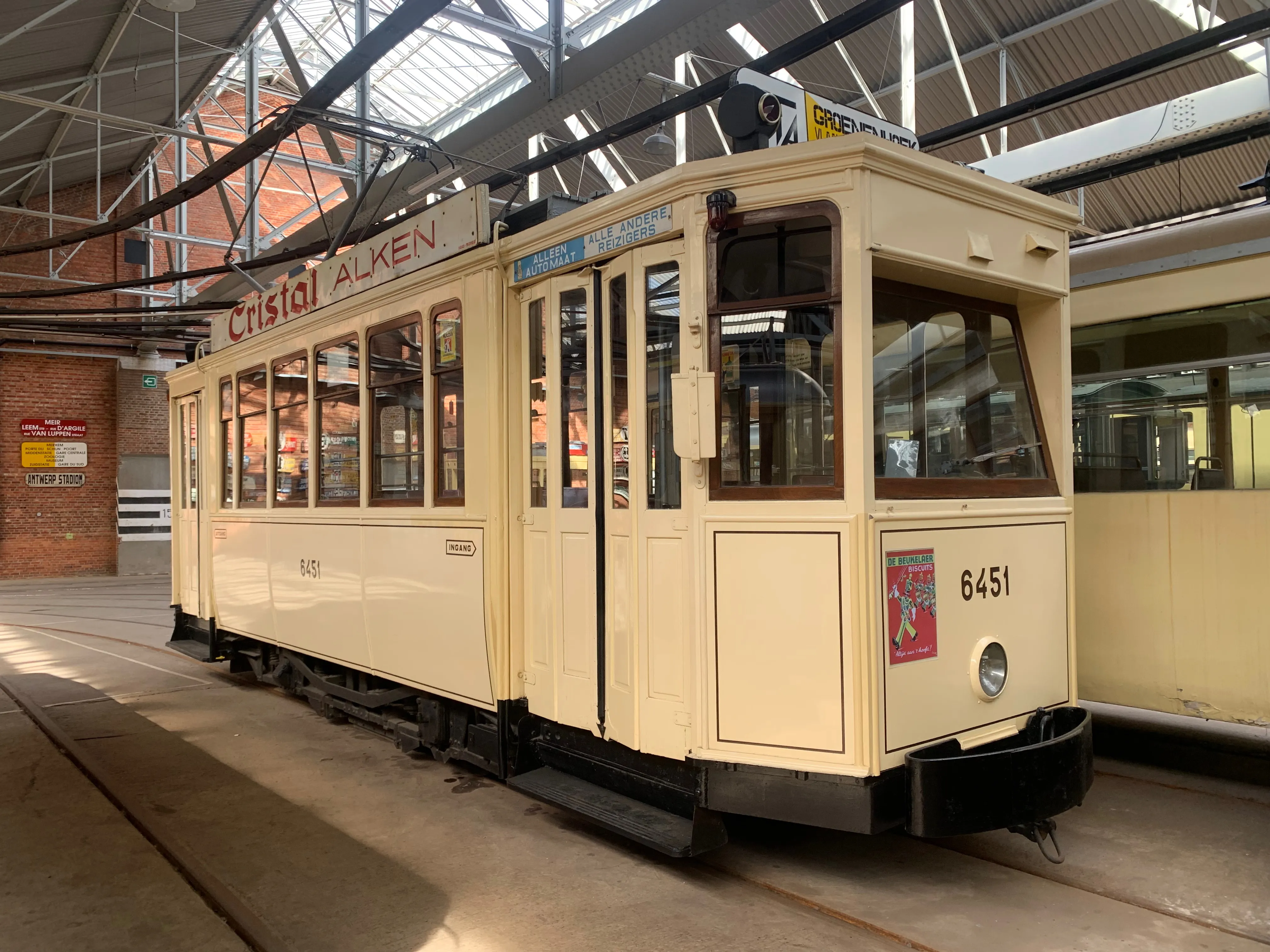 Historyczny tramwaj w kolorze kremowym o numerze 6451 z reklamą Cristal Alken na dachu, wyeksponowany wewnątrz dużej zajezdni o konstrukcji dachowej ze szkła i stali