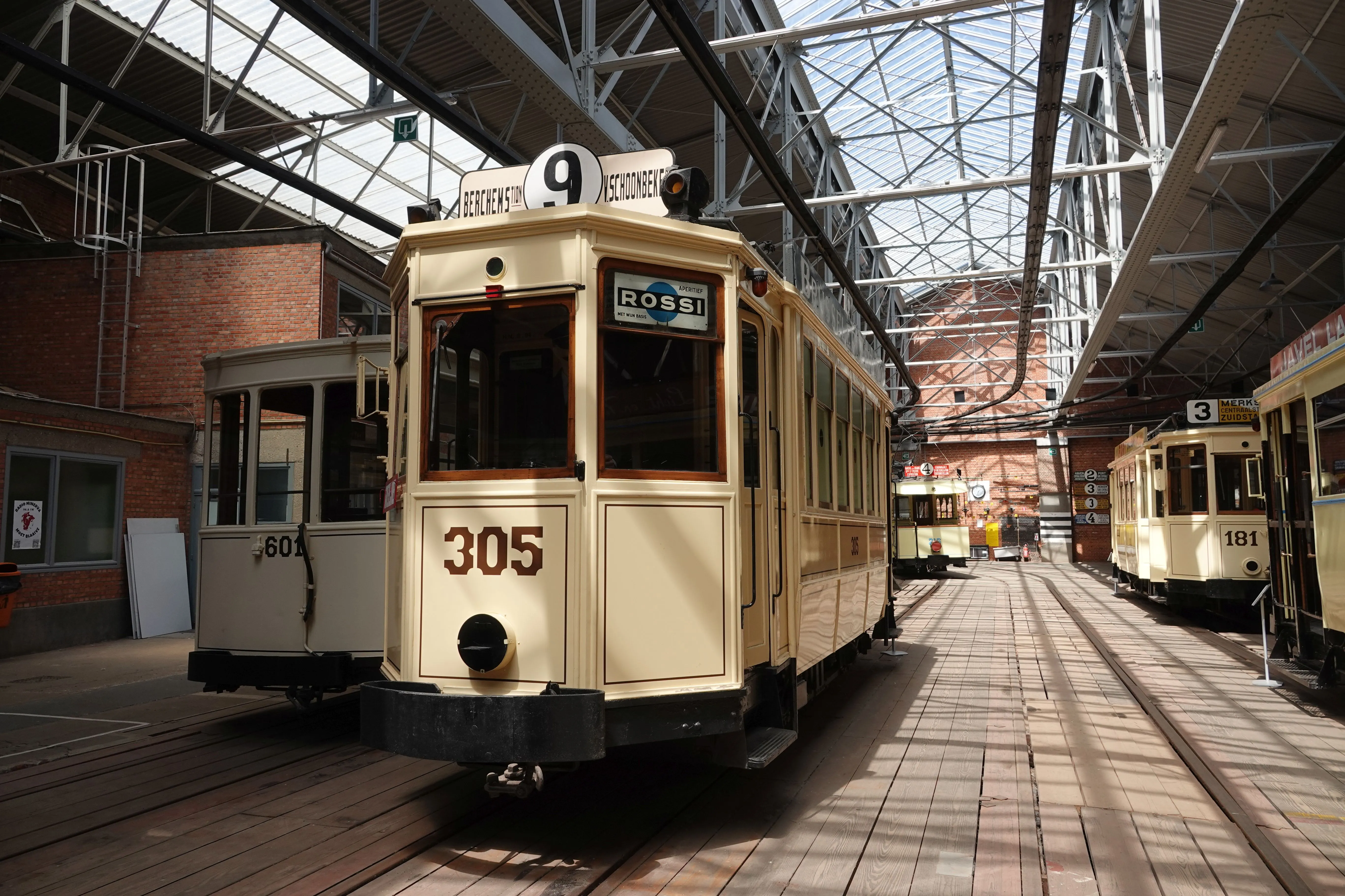 Kremowy zabytkowy tramwaj numer 305 z tablicą kierunkową 9, prezentowany w dużej zajezdni ze szklanym dachem obok innych zabytkowych tramwajów na torach.