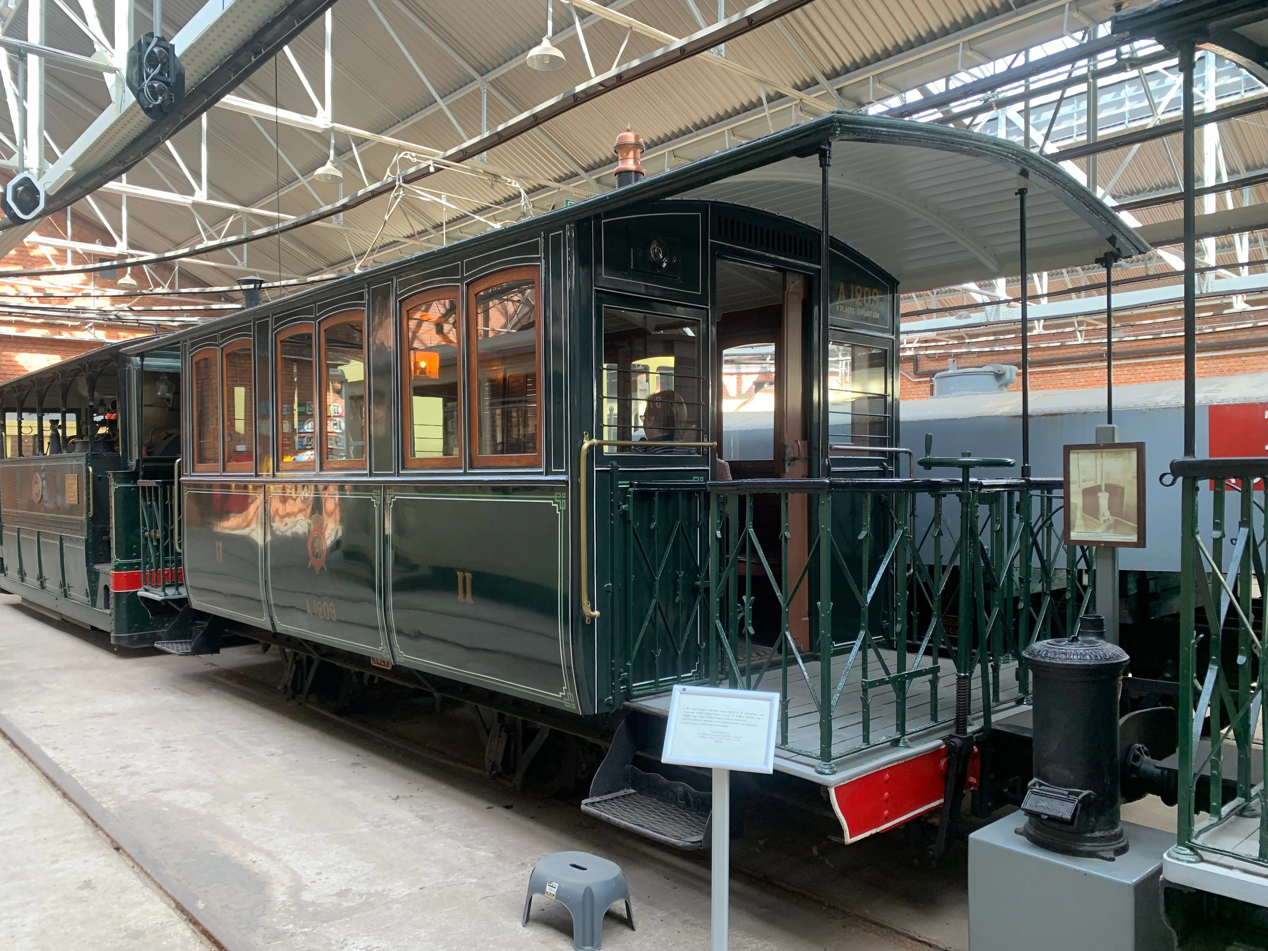Un tranvía histórico de color verde oscuro en exhibición dentro de un depósito de museo, con ventanas de marco de madera y una plataforma trasera abierta con barandillas de metal verde.