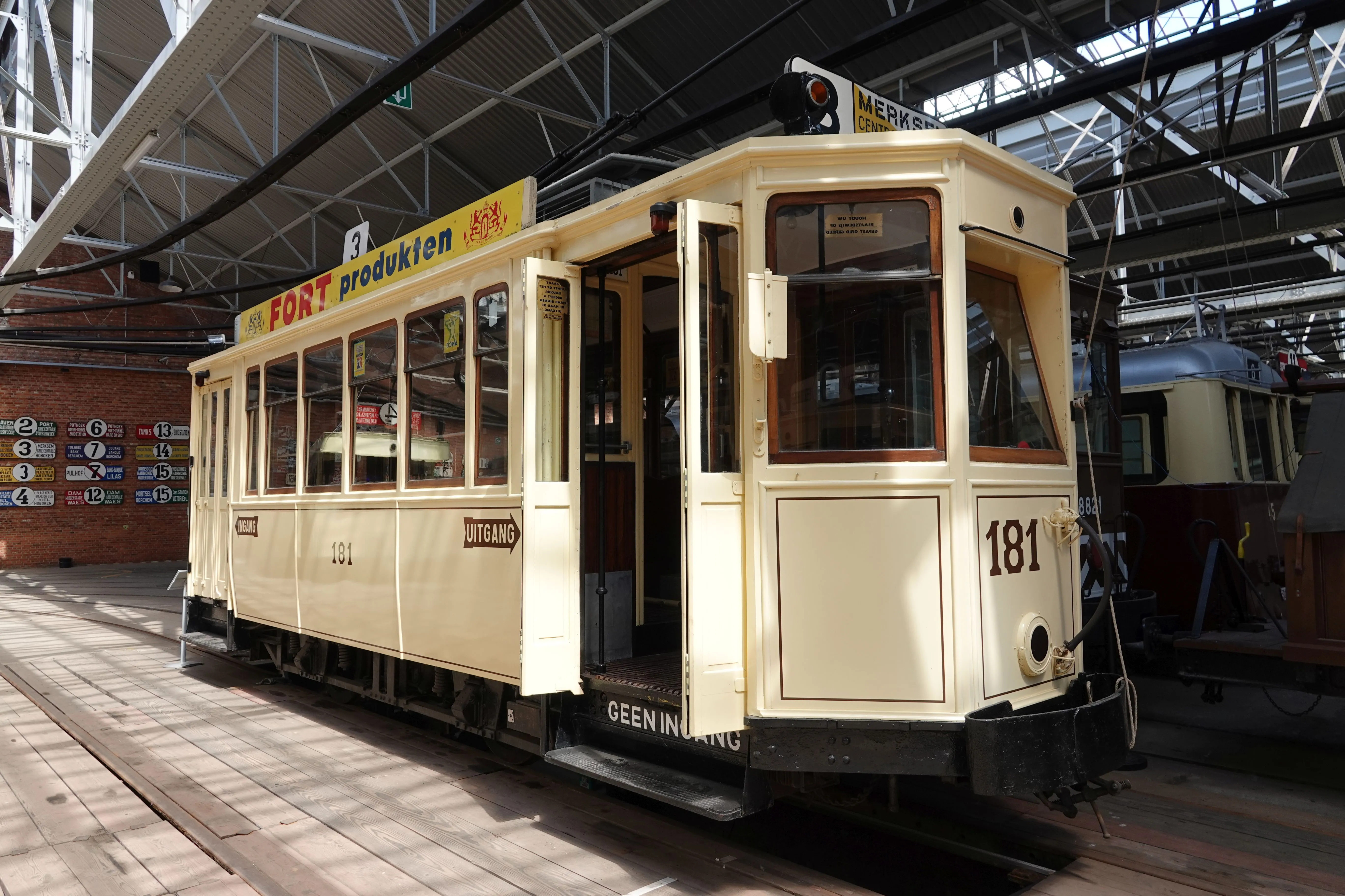 Zabytkowy tramwaj w kolorze kremowym o numerze 181, z żółtą reklamą dachową FORT produkten, zaparkowany wewnątrz muzeum zajezdni tramwajowej VlaTAM, z innymi zabytkowymi tramwajami widocznymi w tle.