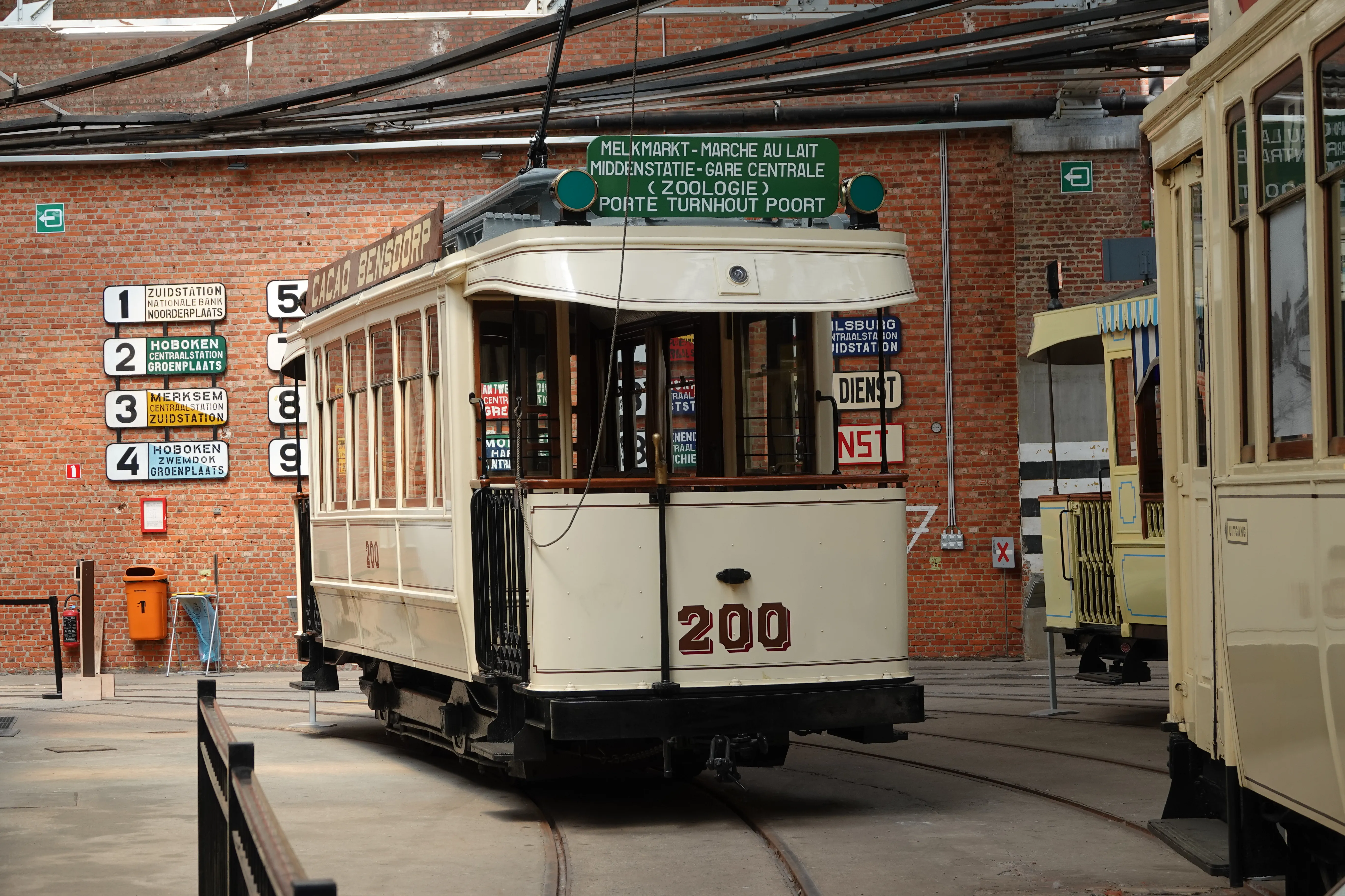 Historyczny tramwaj w kolorze kremowym o numerze 200, wyeksponowany w muzeum zajezdni tramwajowej o ceglanych ścianach, z zieloną tablicą kierunkową na dachu i numerami linii na ścianie za nim.