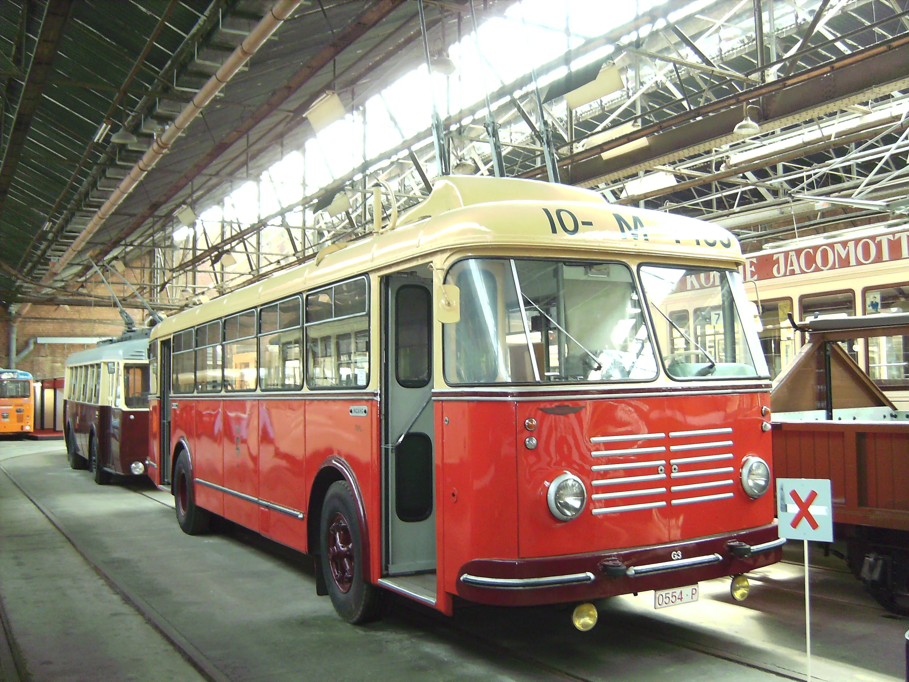 Zabytkowy żyrobus w kolorze czerwono-kremowym o numerze 10 zaparkowany w dużej hali zajezdni, z widocznymi w tle innymi zabytkowymi tramwajami i autobusami.