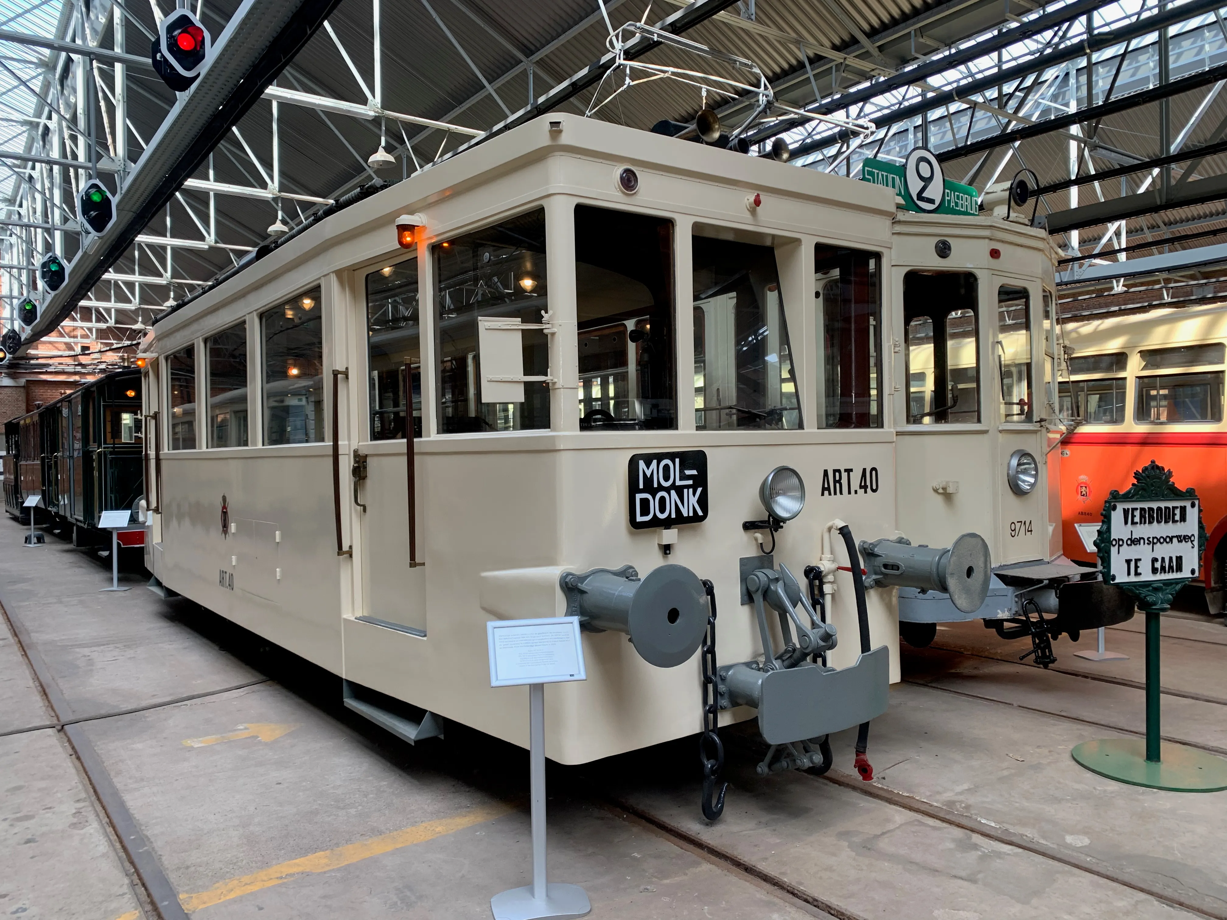 Kremowy zabytkowy tramwaj z napisem 'MOLENBEEK ART.40' wystawiony wewnątrz dużej hali zajezdni ze szklaną konstrukcją dachu i wiszącą sygnalizacją świetlną.
