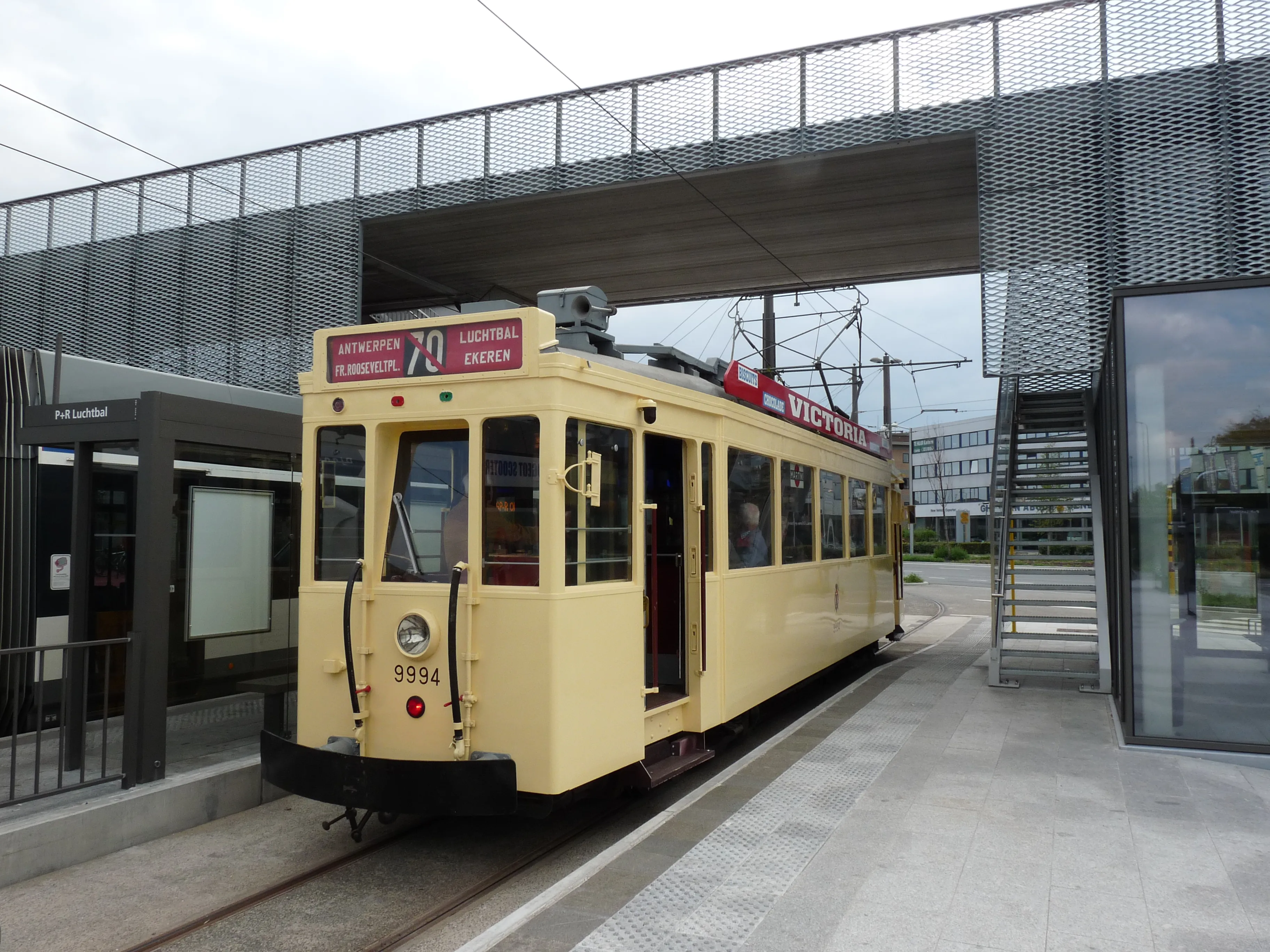 Zabytkowy tramwaj w kolorze kremowym o numerze 20 z czerwoną tablicą kierunkową 'CENTRUM STATION', wyjeżdżający z nowoczesnej zajezdni z metalowej siatki w muzeum Vlaams Tram- en Autobusmuseum.