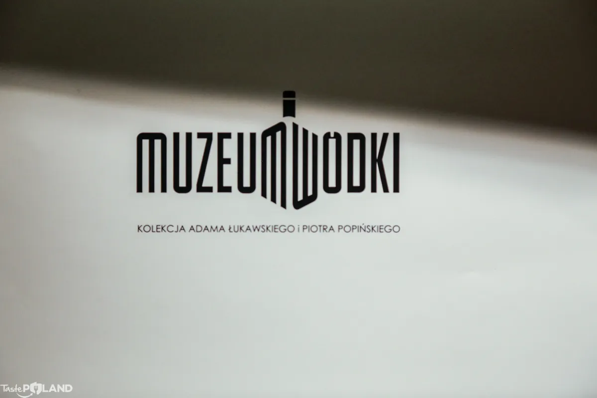 Czarny tekstowy logotyp na białym tle z napisem MUZEUM WODKI i wzorem w kształcie butelki w literze M, a pod nim mniejszy tekst wskazujący na kolekcję Adama Łukawskiego i Piotra Popińskiego.
