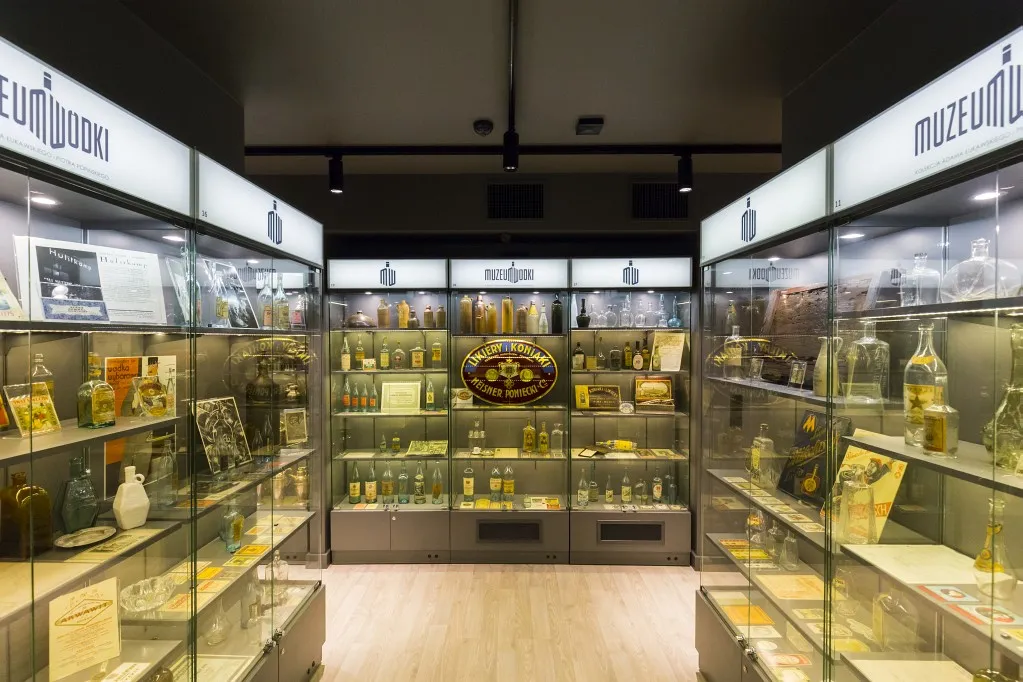 Wnętrze muzeum z szklanymi gablotami wypełnionymi butelkami wódki i pamiątkami, nad nimi podświetlany napis 'MUZEUM WÓDKI'.