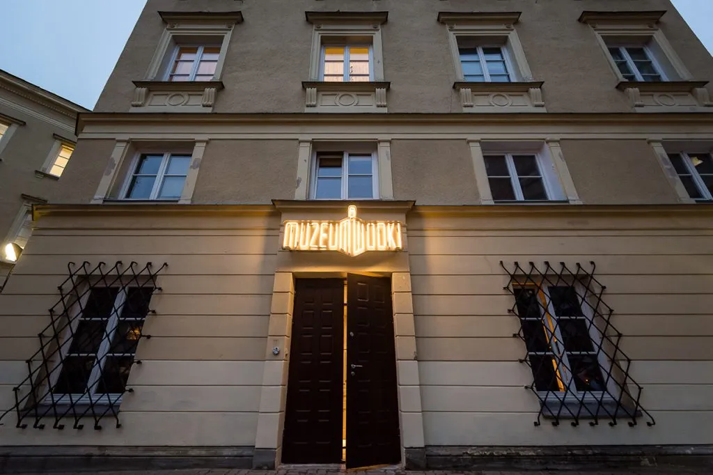 Wejście do Muzeum Wódki z podświetlanym szyldem nad drzwiami, z beżową fasadą i kratami okiennymi z czarnego metalu.