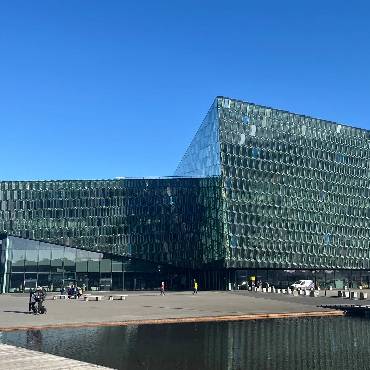 Wide shot of the Harpa building's exterior featuring a large geometric glass facade under a clear blue sky, with a waterfront plaza and people walking in the foreground.