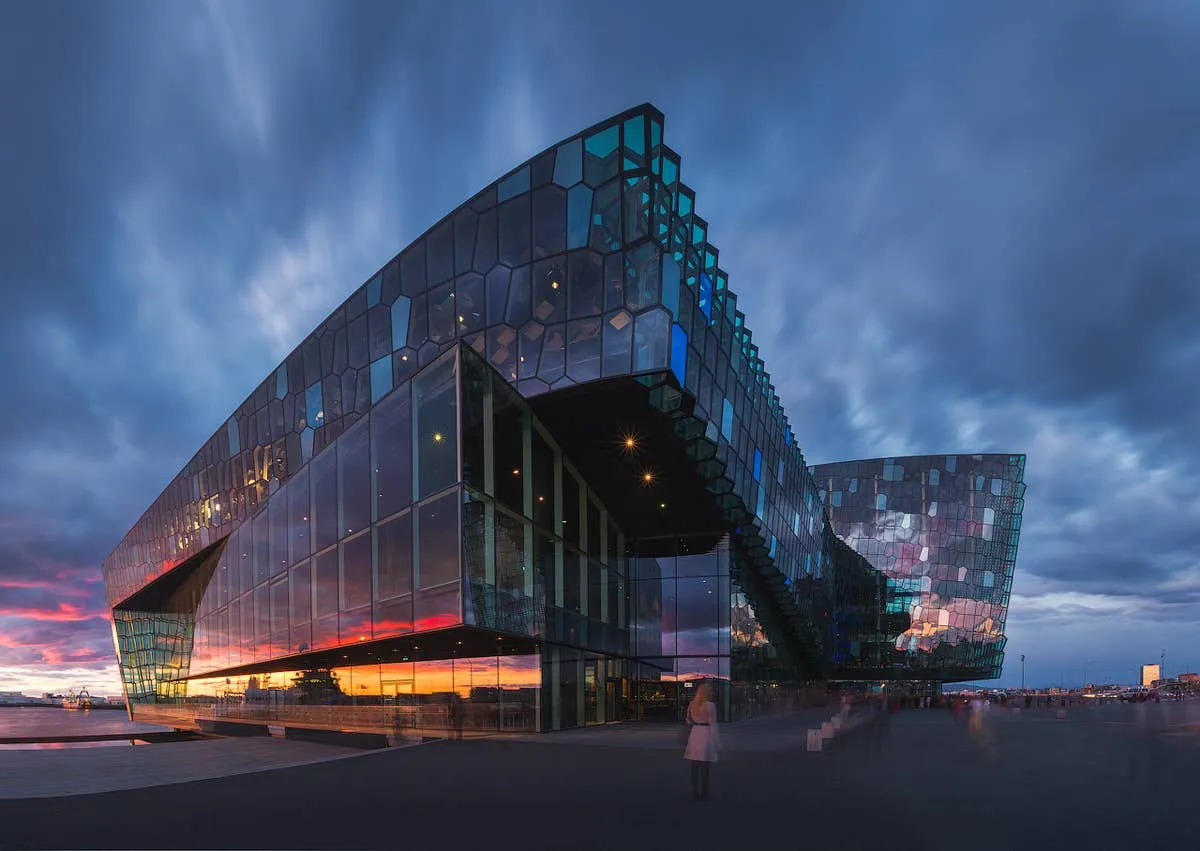 Angular glass-facade building with geometric honeycomb panels glowing under a dramatic blue and orange twilight sky, with a few people walking on the waterfront plaza.