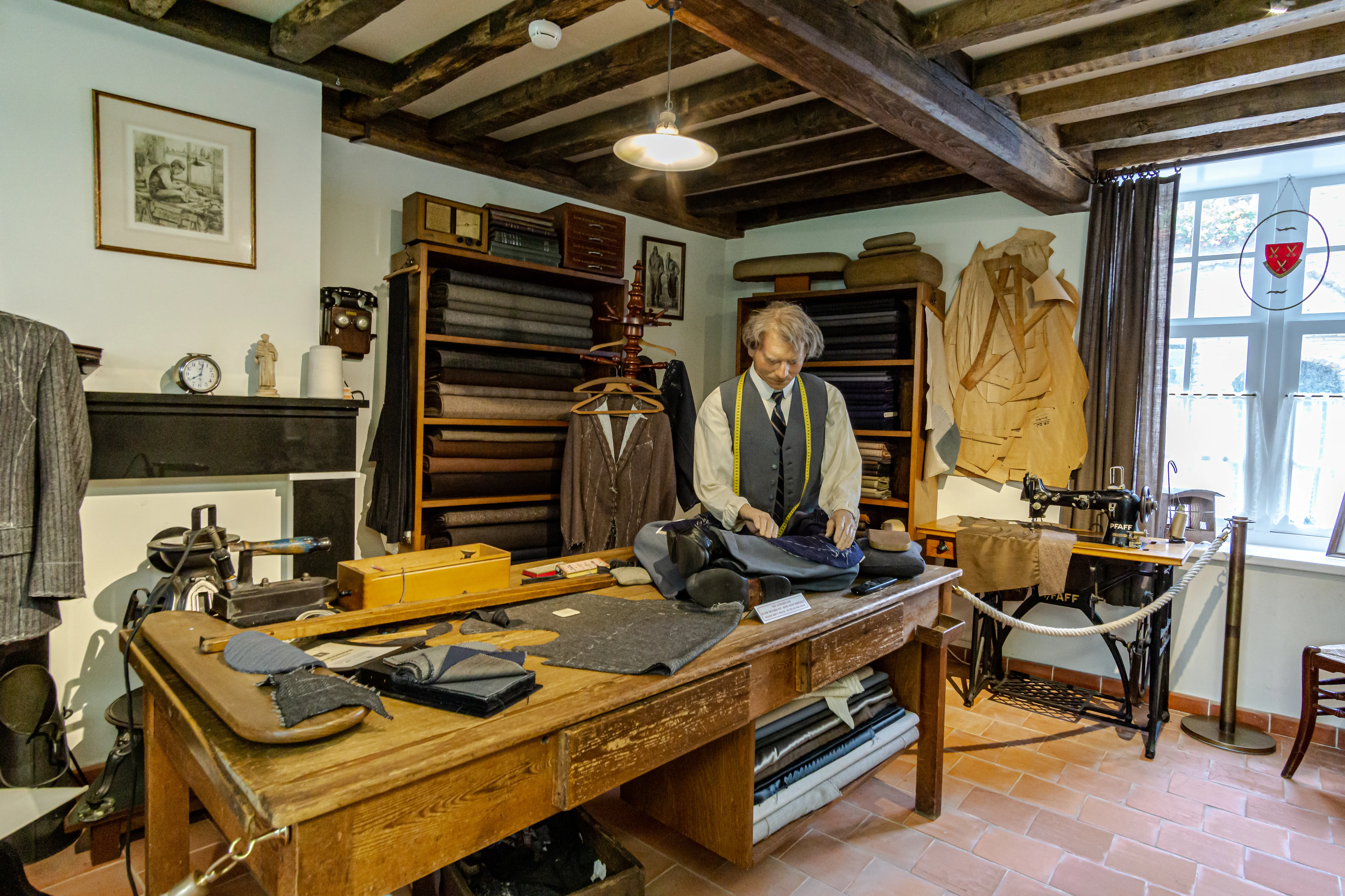 Interior recreado de un taller de sastre con un maniquí vestido de sastre trabajando en una gran mesa de madera, rodeado de telas, herramientas de costura, estantes con rollos de tela y una máquina de coser bajo vigas de madera vistas en el techo.