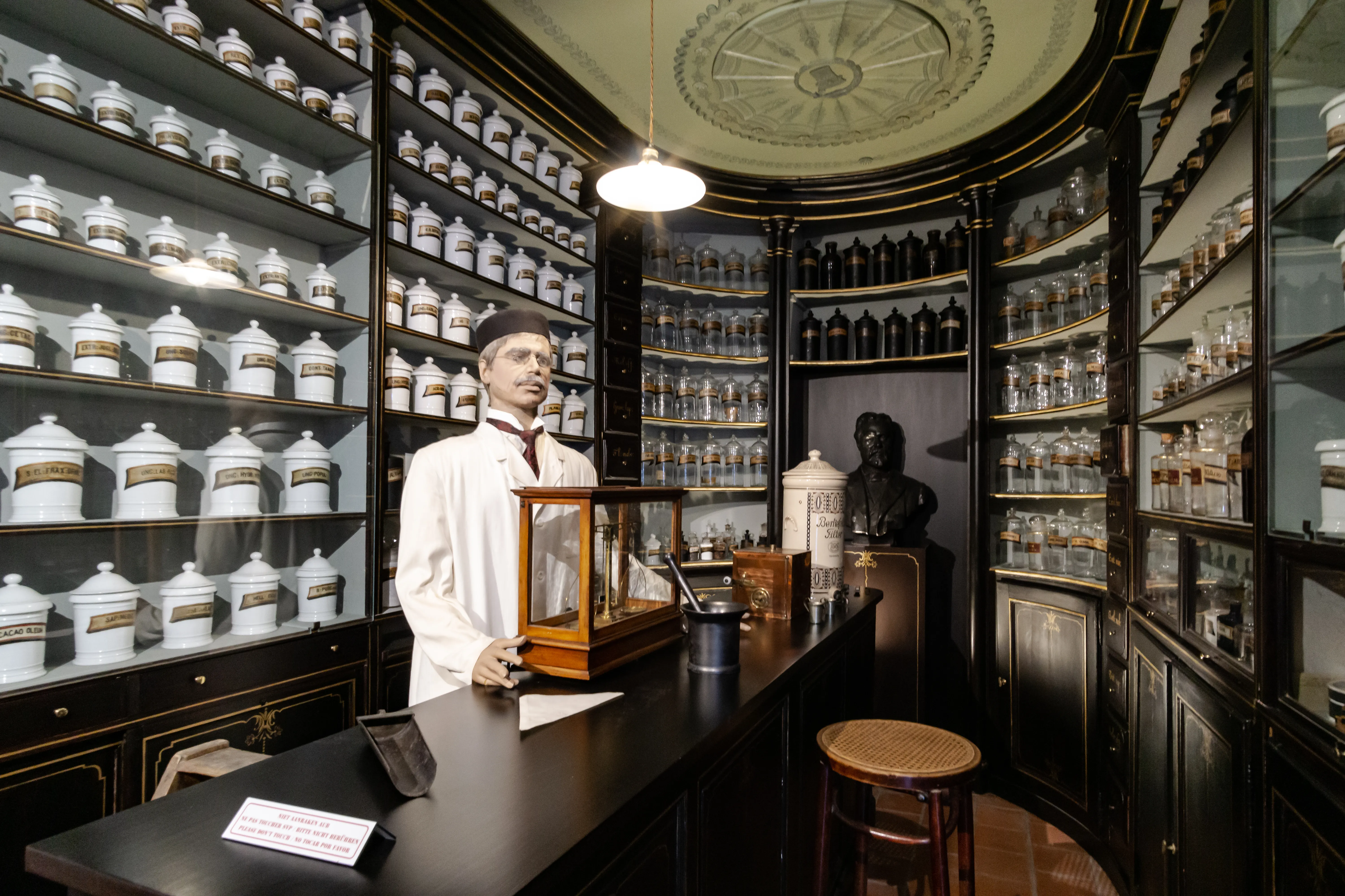 Interior de una farmacia histórica recreada con estanterías de suelo a techo llenas de frascos de cerámica blanca y botellas de vidrio, un maniquí con bata de farmacéutico blanca detrás de un mostrador de madera oscura, un busto de bronce y un medallón ornamentado en el techo.