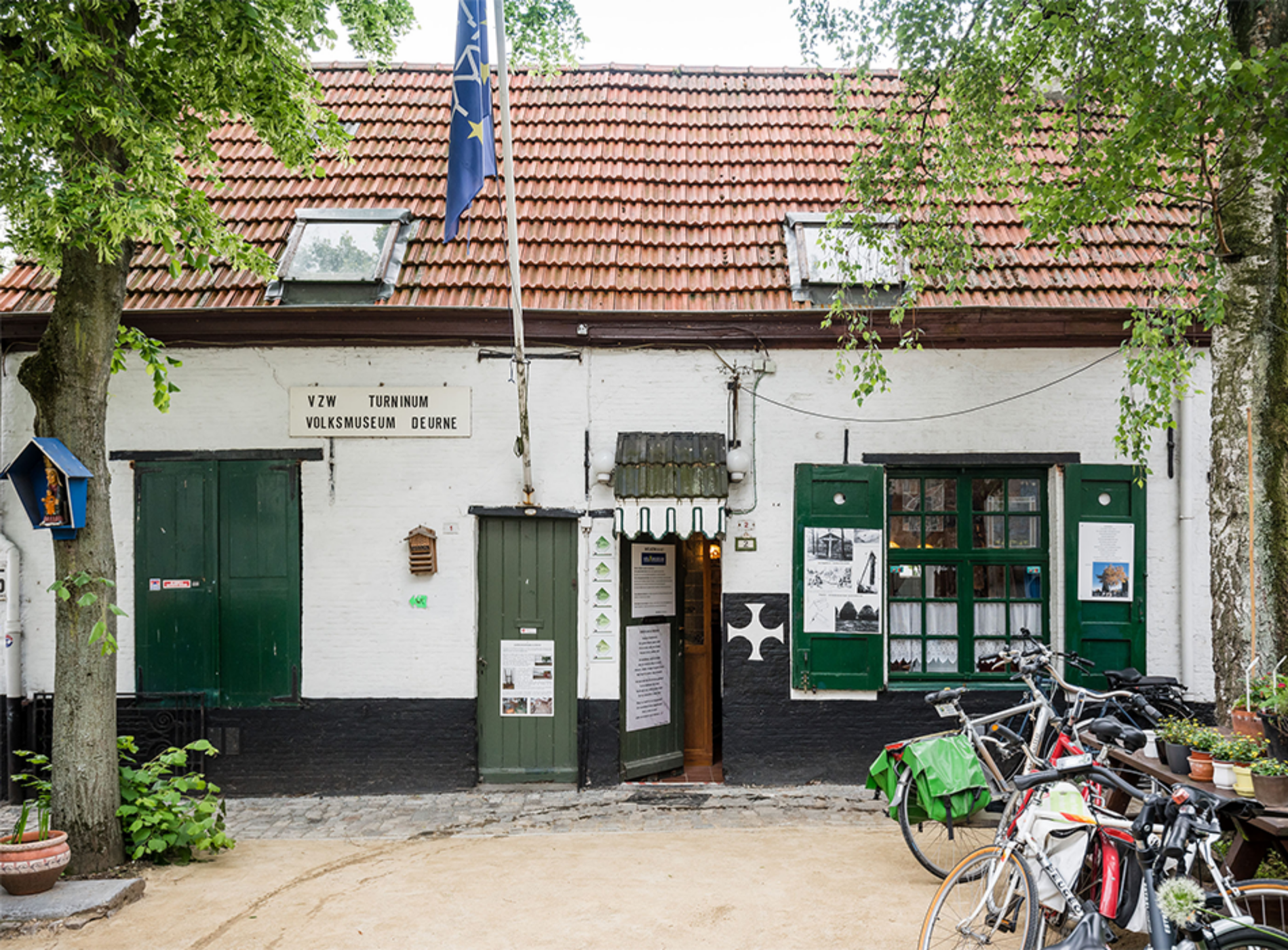 Biały budynek z czerwonym dachem z dachówek, zielonymi drzwiami i okiennicami oraz szyldem z napisem V.H.M. Tonnen Volksmuseum Deurne, przed którym zaparkowane są rowery