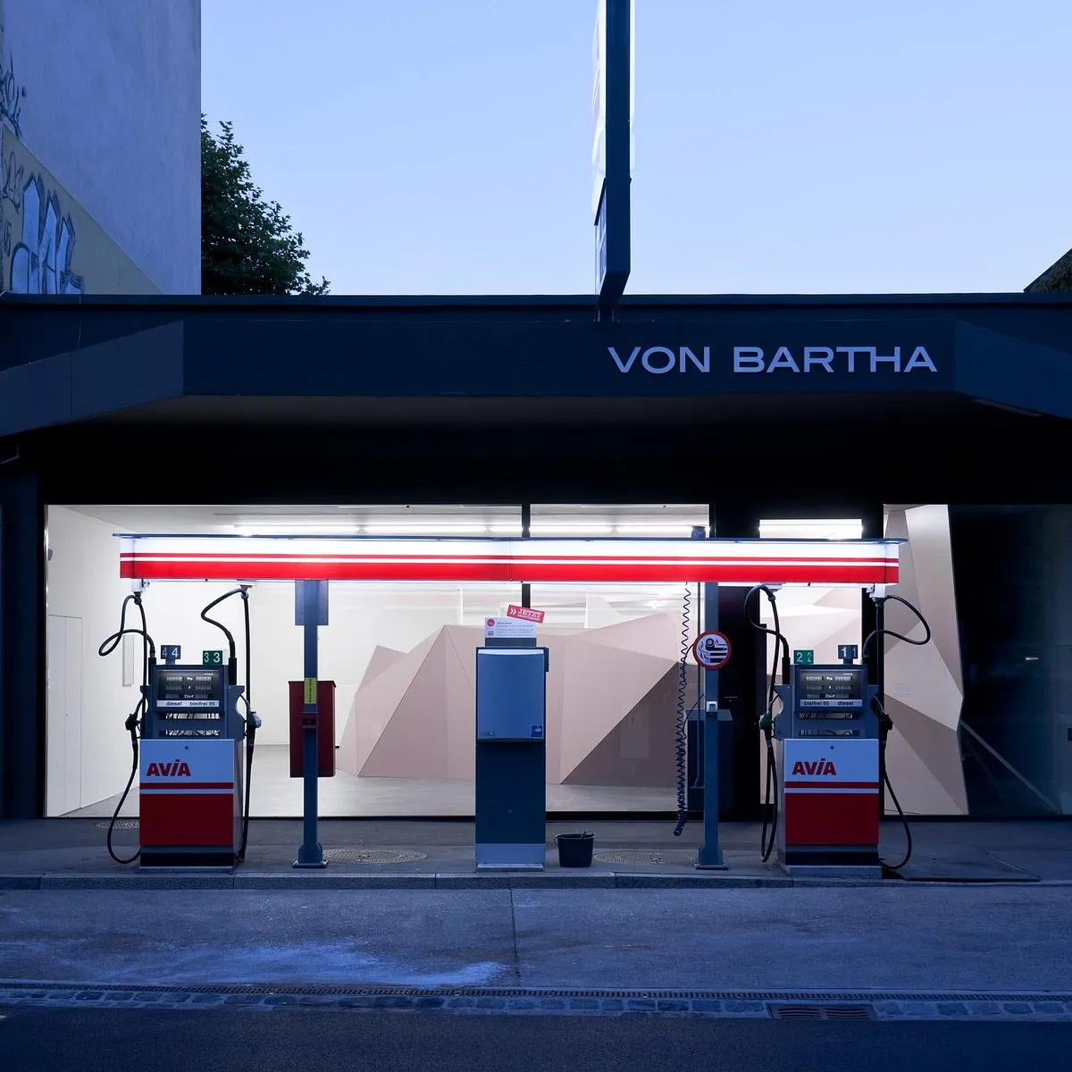 Exterior view of von Bartha gallery showing the dark facade with VON BARTHA sign above an open garage door revealing an art installation with vintage AVIA gas pumps under a red neon light strip