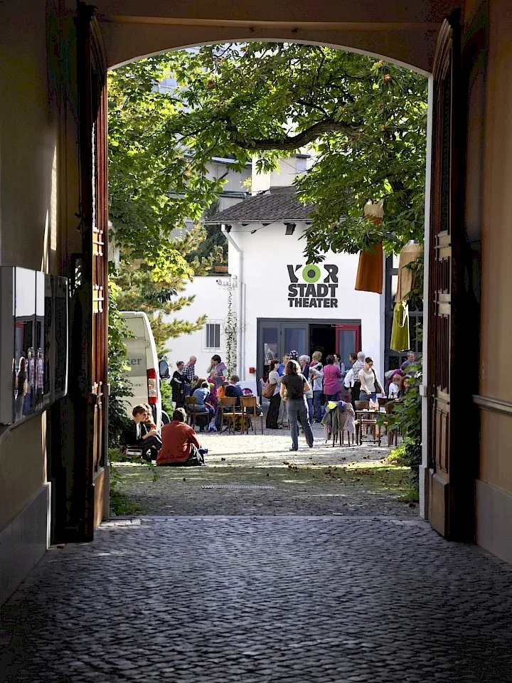 Vista a través de un portal arqueado hacia un patio soleado donde la gente se reúne frente a un edificio blanco con el letrero de Vorstadt Theater, con un camino de adoquines en primer plano