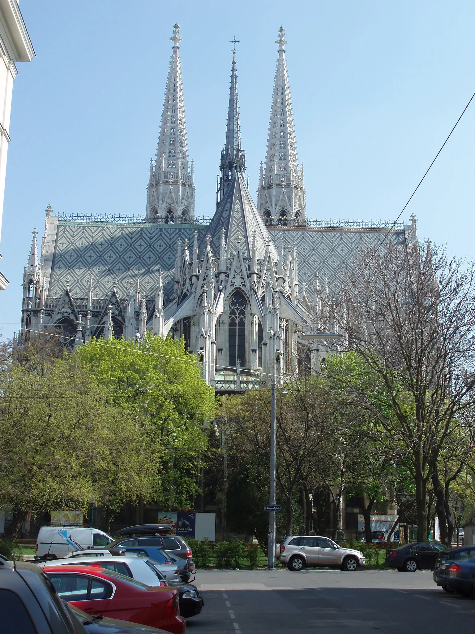 Neo-Gothic church with twin spires and ornate stonework, surrounded by trees and parked cars