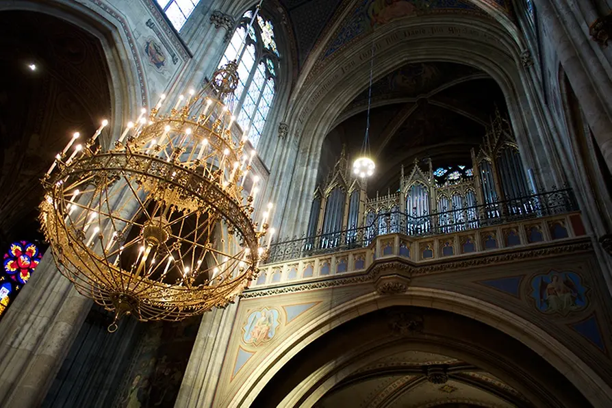 Interior of a church with a large golden chandelier hanging from a high vaulted ceiling and a pipe organ on a balcony