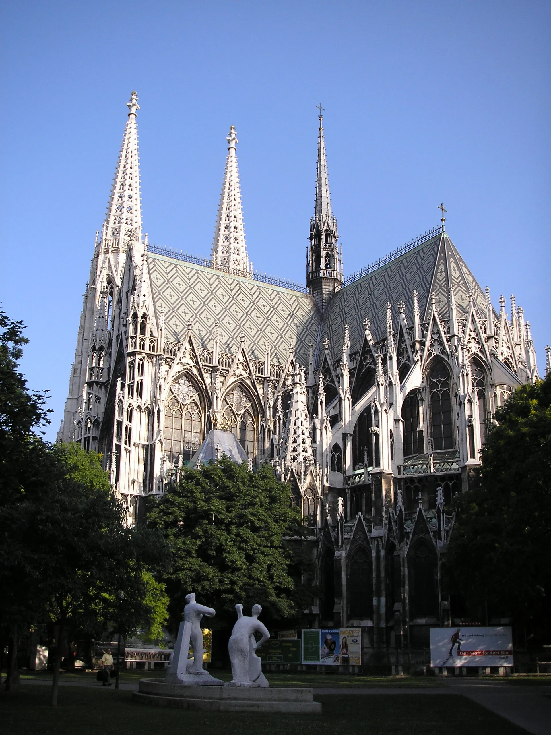 Neo-Gothic church with twin spires and detailed stonework under a clear blue sky, surrounded by trees and a white sculpture in the foreground