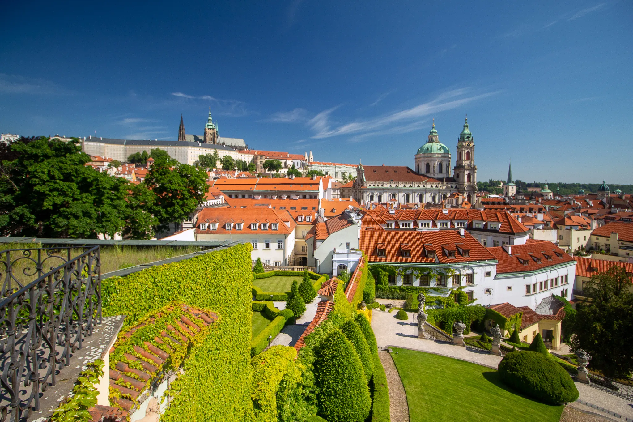 Vrtba Garden met groene terrasvormige hagen en uitzicht op de historische daken en kerktorens van Praag onder een blauwe hemel