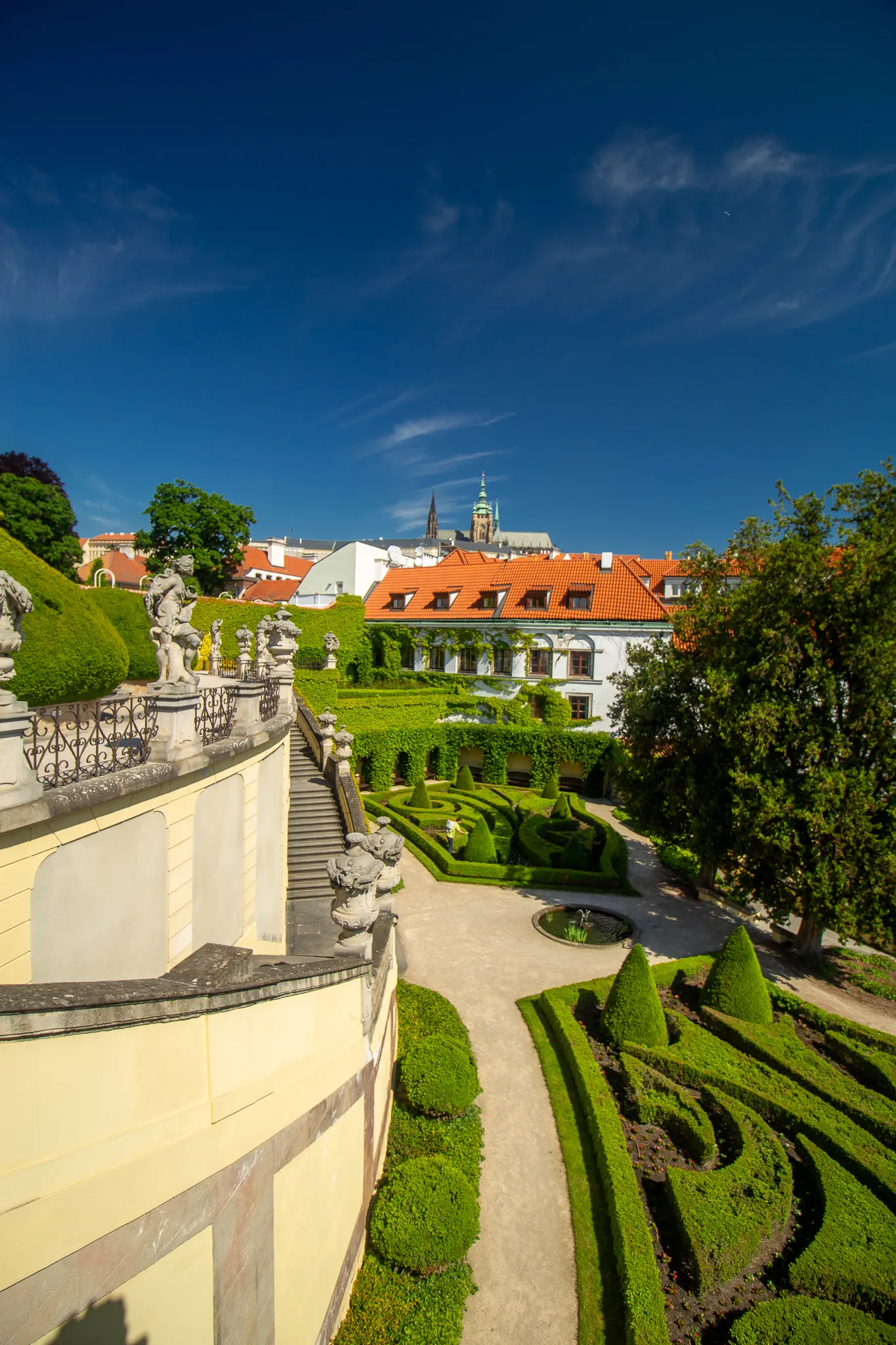 Panoramisch uitzicht op de Vrtba-tuin met verzorgde groene hagen, stenen paden en een helderblauwe hemel