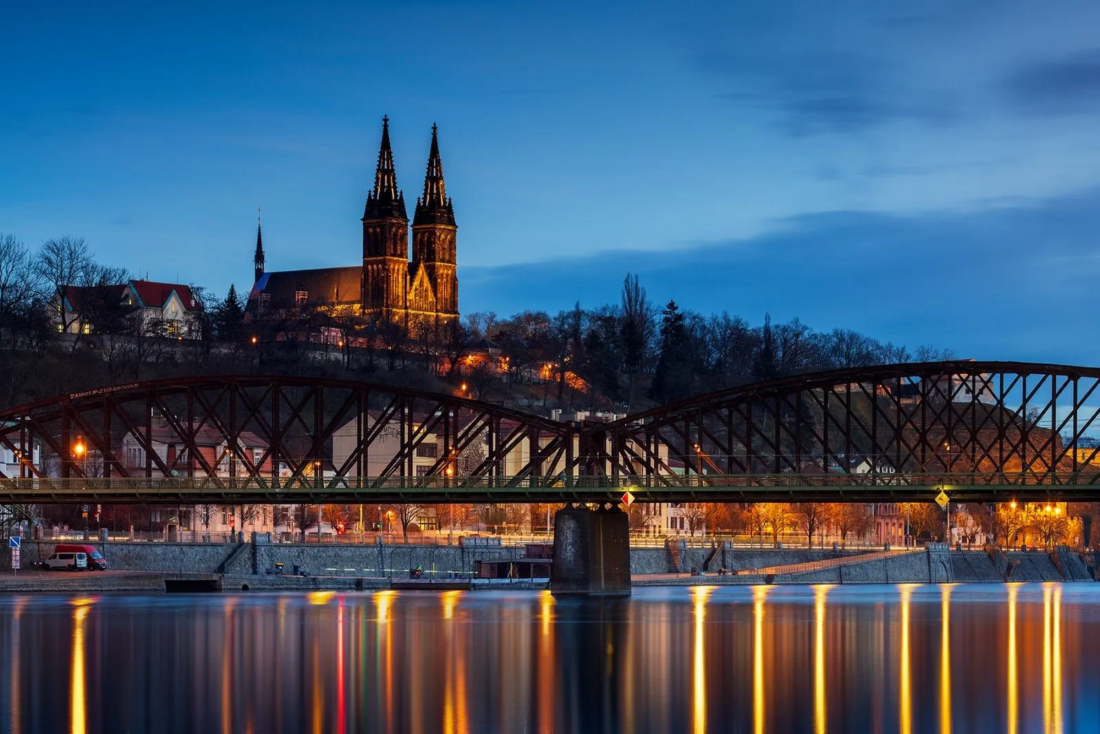 Basílica iluminada con dos campanarios en una colina con vistas a un río con un puente y reflejos al anochecer