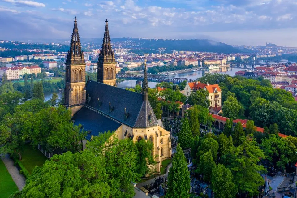 Vista aérea de una gran iglesia con campanarios gemelos rodeada de árboles verdes, con una ciudad y un río al fondo.