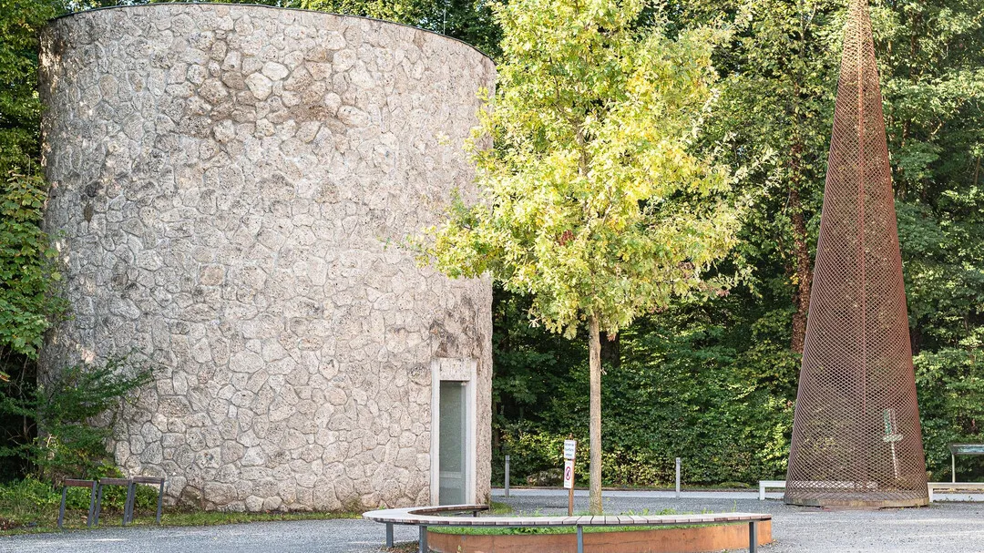 Una gran estructura cilíndrica de piedra se alza junto a una alta escultura metálica cónica oxidada y un árbol joven en un entorno artístico público pavimentado.