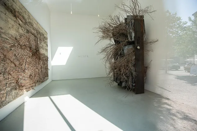 Interior de una galería con una gran obra de arte de textura marrón en la pared de la izquierda y una alta instalación escultórica de ramas y paneles de madera oscura a la derecha, con luz natural proveniente de una claraboya.