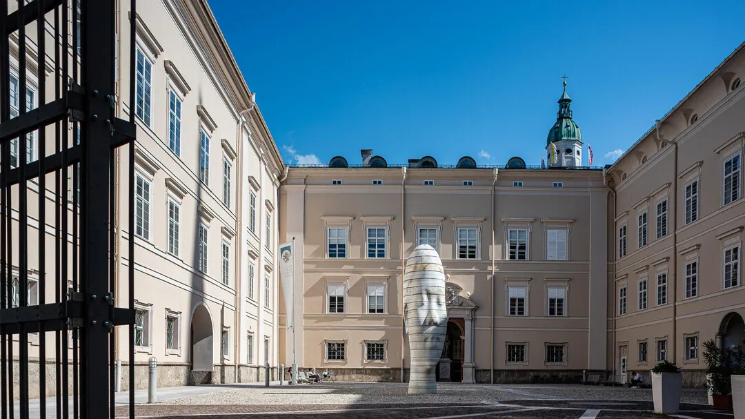 Gran instalación escultórica de una cabeza blanca situada en el centro de un patio histórico de Salzburgo, rodeada de edificios clásicos de color crema con la torre de una iglesia de cúpula verde al fondo.