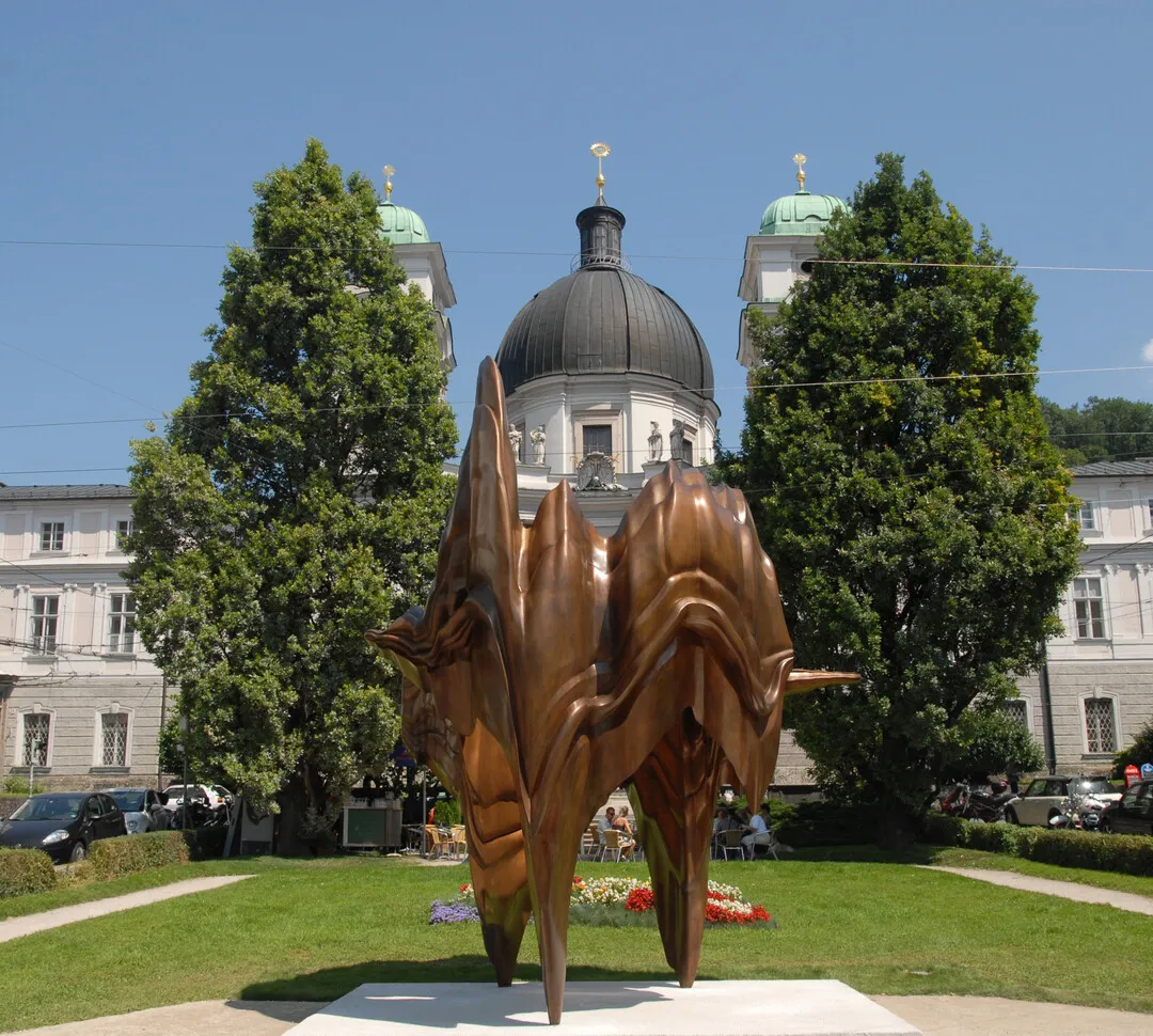 Gran escultura abstracta de bronce sobre una base blanca en un parque verde, con una iglesia barroca con cúpula y árboles al fondo